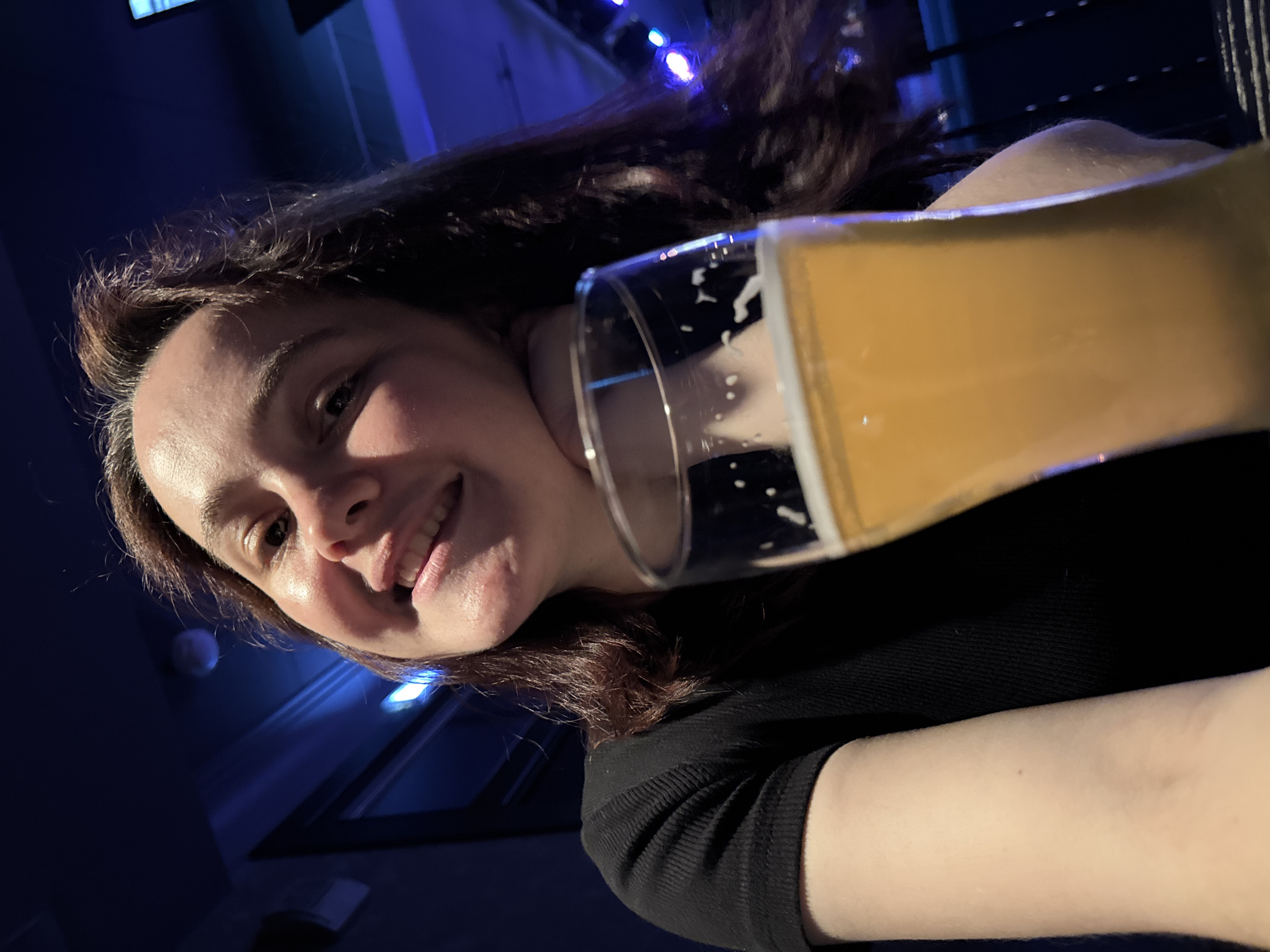 In the foreground of the image, a glass of golden-yellow liquid, likely beer, is held up, obscuring part of the scene. The glass has white foam clinging to its rim and sides. Behind the glass, a young woman with long, dark brown hair is smiling. Her right hand is propped under her chin, and she is wearing a black, ribbed t-shirt. The background is dimly lit with blue ambient light, suggesting an indoor setting like a bar or restaurant. There are hints of other patrons or a stage with lights in the blurred background. The image was taken in Izhevsk, Russia.