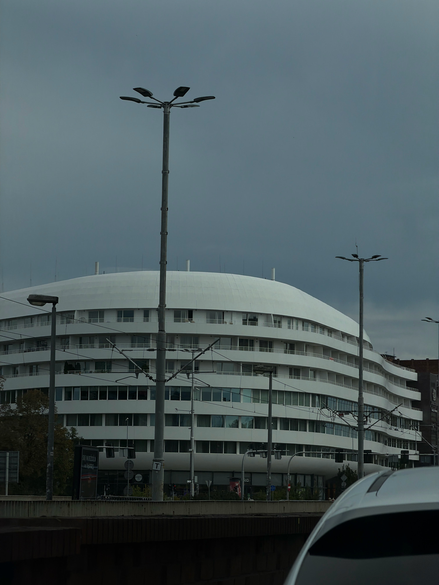 The image shows the white, curved facade of a modern, multi-story building in Wrocław, Poland, under a cloudy sky. Several tall, grey lampposts with multiple lights are visible in the foreground and middle ground, along with overhead electrical wires that appear to be for a tram system. The building has numerous windows and balconies wrapping around its curved structure. In the lower right corner, the white roof and rear section of a modern car are visible, partially obscuring the view of the street and sidewalk below the building. A dark, textured surface, possibly a bridge or elevated walkway, occupies the bottom of the frame. Signs and traffic lights are visible at street level. Trees with sparse autumn foliage are present on the left side.