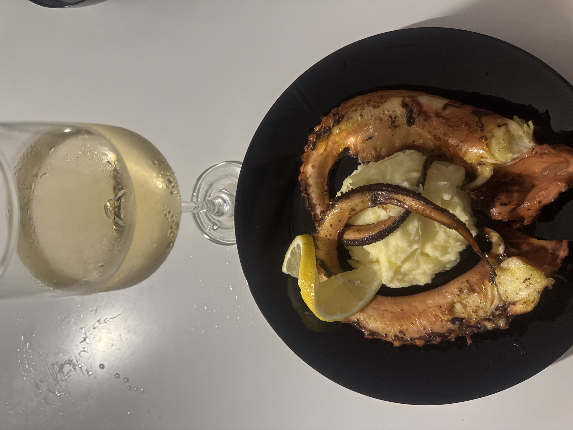 A top-down shot captures a meal consisting of seafood and a beverage on a light-colored, reflective surface. A round black plate occupies the lower portion of the frame, holding two large, cooked octopus tentacles or legs, characterized by their reddish-brown, slightly charred exteriors. A mound of mashed potatoes is centered between the octopus pieces, garnished with two slices of yellow lemon. Above the plate, a stemmed wine glass holds a light, clear yellow liquid, likely white wine, exhibiting condensation on its exterior and small bubbles within the liquid, indicating it is chilled. Water droplets and faint streaks are visible on the white surface, particularly around the glass and the upper left area of the image. The overall setting is indoors. The location of Varna, Bulgaria cannot be determined from the visual information provided.