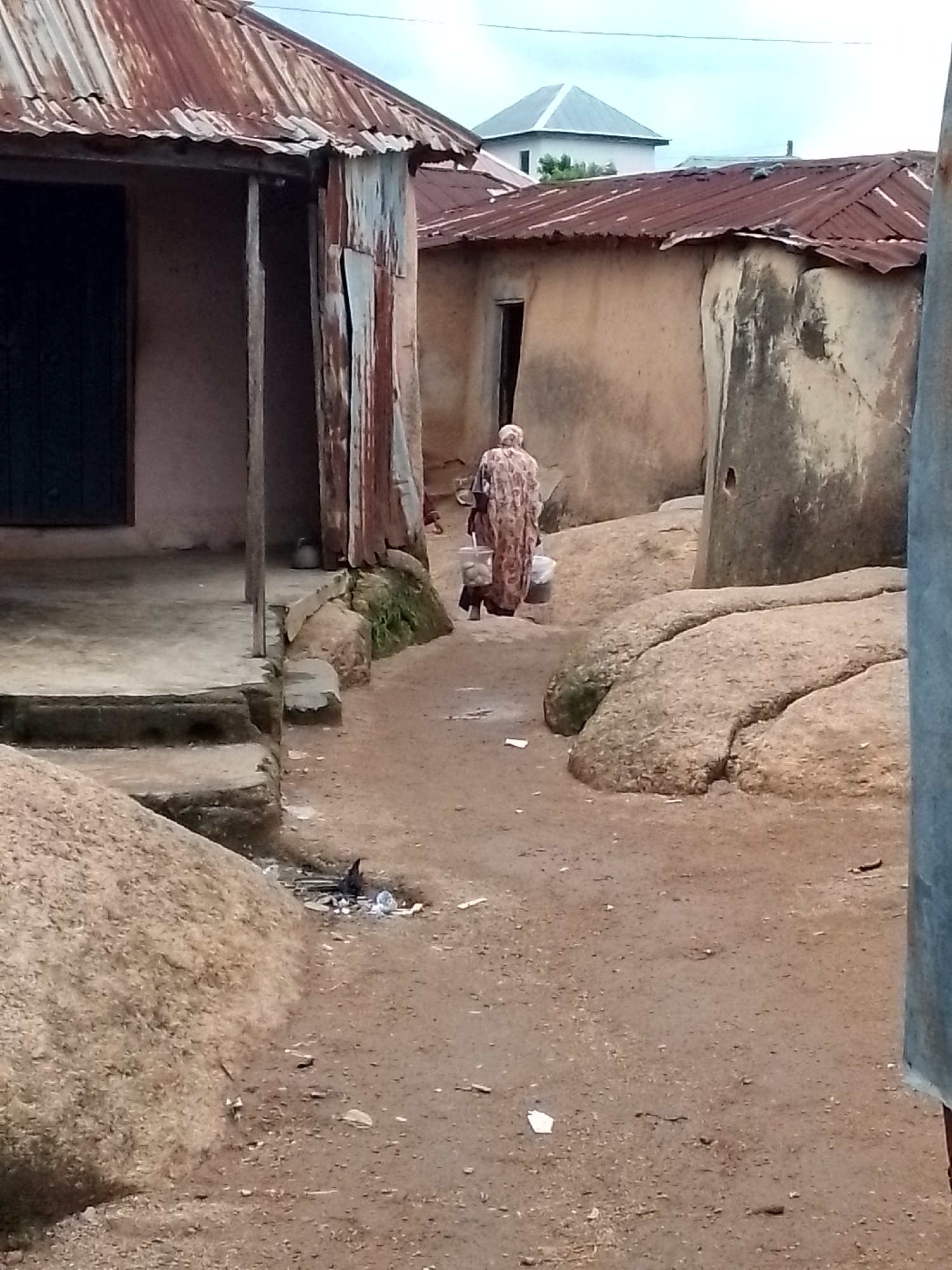 A person wearing a patterned garment walks away from the camera down a dirt path between buildings. The person is carrying buckets. The buildings have corrugated metal roofs and mud or concrete walls. Large rocks are visible in the foreground and to the right of the path. The ground is dry and dusty, with some scattered debris. The sky is overcast.