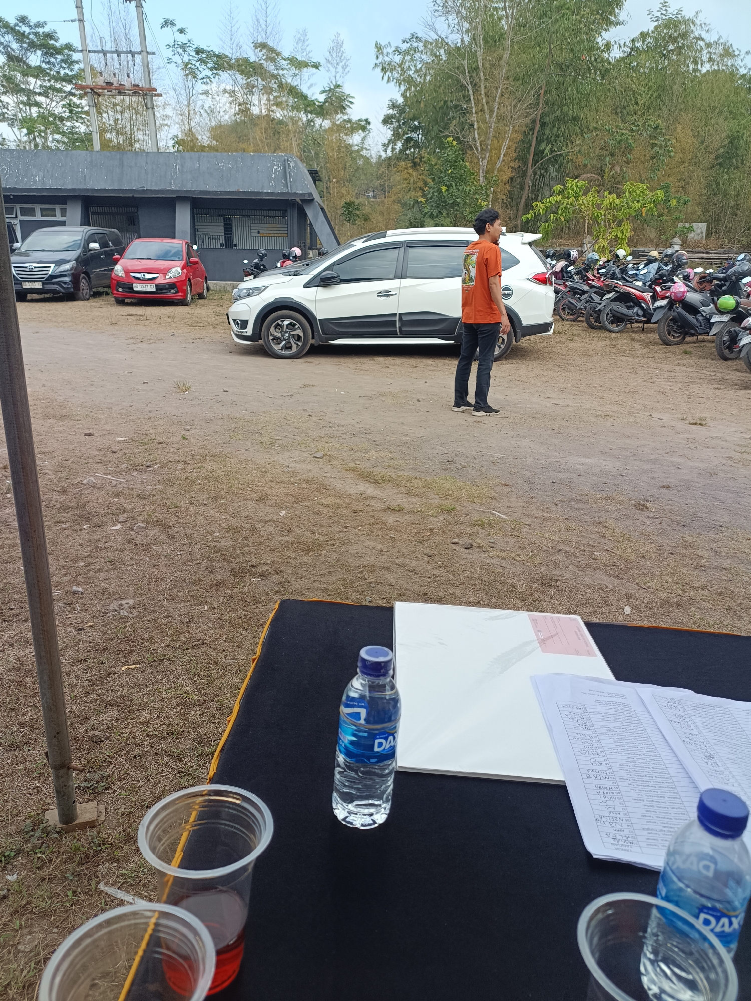 A full shot captures an outdoor scene with a parking lot in front of a low-rise building. In the foreground, a black table is set up with two clear plastic cups containing reddish liquid, a bottle of DAX water, and some paperwork. To the right of the table, a person stands facing left, wearing an orange shirt and black pants. Behind them, a white SUV is parked, and a row of motorcycles is visible further back. In the background, the building has a gray facade, and a power line pole stands to its left. The ground is a dirt and gravel surface with patches of dry grass. The scene is likely in Hargobinangun, Indonesia, given the context.