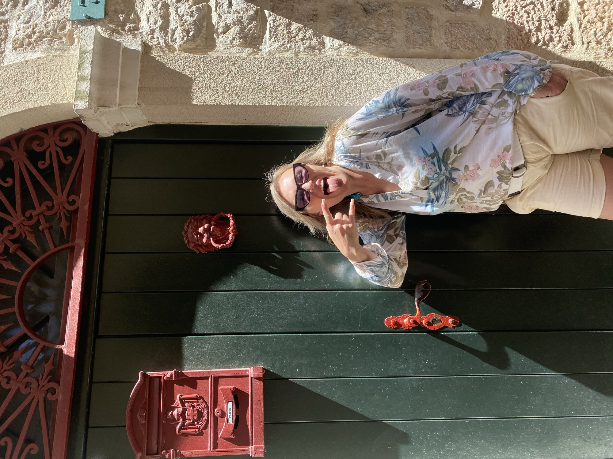 The image shows a fair-skinned, blonde-haired woman with sunglasses standing sideways in front of a green door and stone wall. The woman is wearing a white floral shirt and beige shorts and is making a "rock on" gesture with her hand while sticking out her tongue. The door is dark green with horizontal slats and is decorated with a red metal door knocker in the shape of a lion's head, a red metal mail slot, and a red metal handle. The wall is made of light-colored stone. Based on the architectural style and the presence of signage in the Cyrillic alphabet, this image likely was taken in Budva, Montenegro.