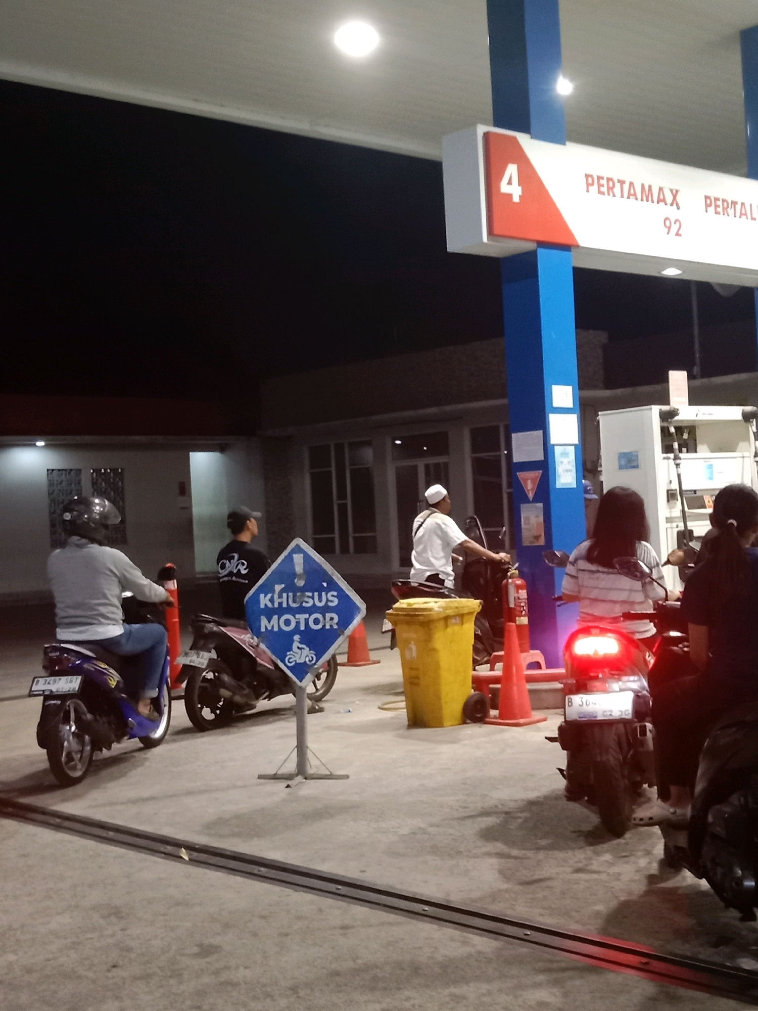 This is a photograph of a gas station in South Tangerang, Indonesia, at night. The gas station has a blue and white color scheme. There are several people on motorcycles, appearing to be waiting to refuel or having just refueled. One sign reads "KHUSUS MOTOR", which means "For Motorcycles Only". Another sign indicates "PERTAMAX 92", a type of fuel. There is a yellow trash can and several traffic cones present. The lighting suggests it is evening or night.