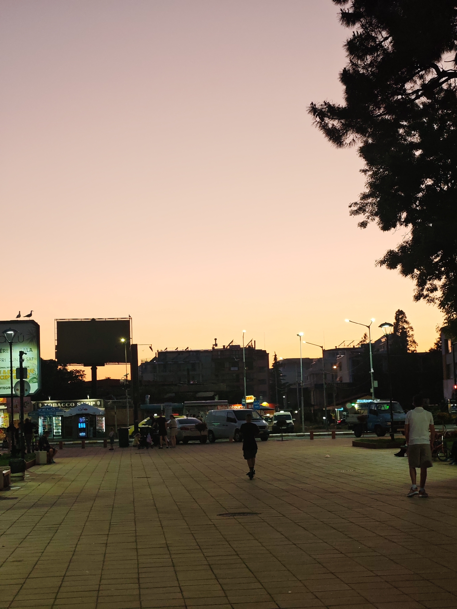 The image depicts a square in what appears to be a city at dusk. The sky is a gradient of orange and pink. Silhouetted against the skyline are buildings, streetlights, and a large advertising billboard. A mature tree occupies the upper right corner of the frame, its branches casting a dark shadow.
The square is paved with light-colored rectangular tiles. Several people are present: a figure is running away from the camera, and another person is walking away from the camera in the same direction. A group of people is standing or sitting near what appears to be a tobacco shop.
Various vehicles are visible on the street beyond the square, including cars and vans. There are also signs indicating a taxi service. The scene is lit by streetlights and the ambient light of the sunset. The overall environment suggests a public space in an urban setting in Nesebar, Bulgaria.