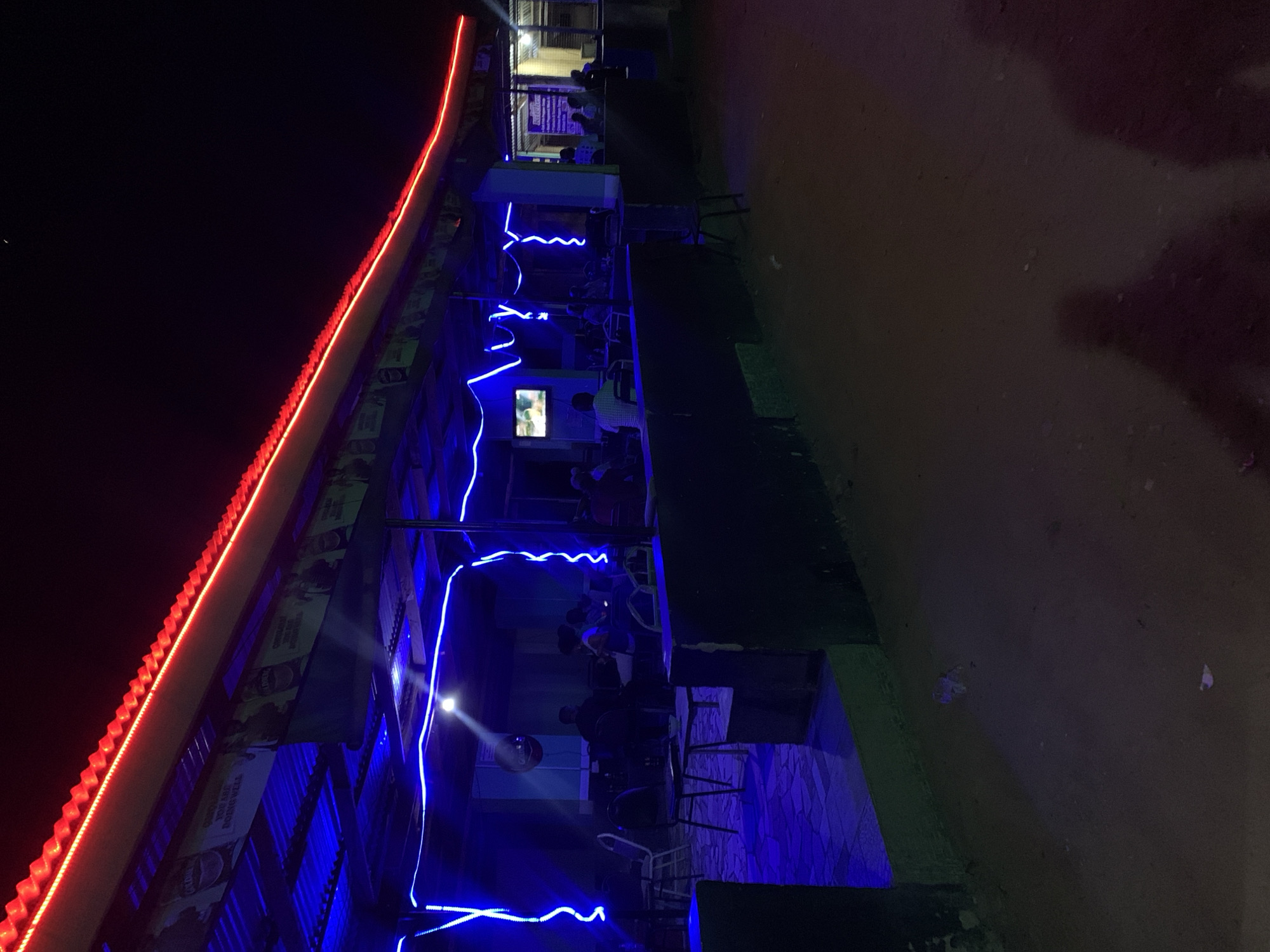 The image shows an outdoor establishment in New Stadium, Nigeria, at night. The building's roof is adorned with red LED lights. Blue LED lights are strung along the building's front, illuminating the seating area. People are seated at tables, presumably customers. A television screen is visible on the building's facade. The surrounding environment appears to be a dirt or unpaved area. Chairs and tables are scattered across the pavement of the establishment.