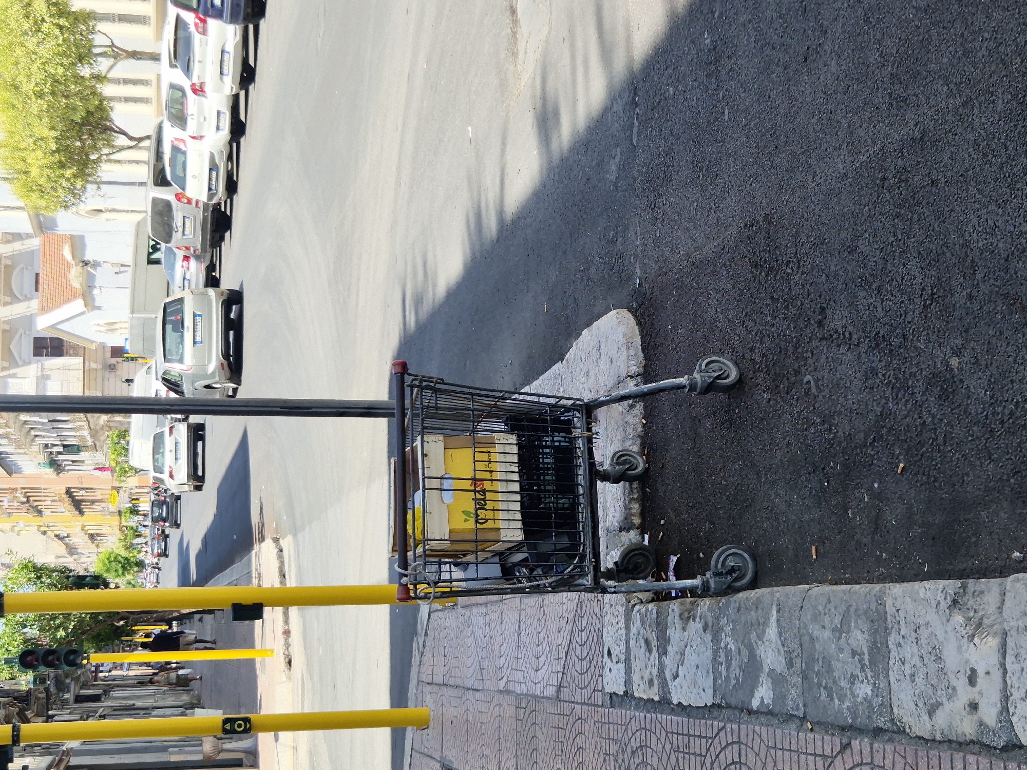 The image depicts a street scene in Taranto, Italy. In the foreground, a metal shopping cart filled with cardboard boxes is parked on the sidewalk next to the street. The cart is black with red handles and has four wheels, two of which are visible and appear slightly worn. The boxes inside the cart are of varying sizes and some have yellow labeling with green text. The sidewalk is paved with patterned tiles and bordered by a concrete curb. The street is asphalt and appears to be a one-way road with cars parked on the right side. Yellow traffic signal poles with traffic lights are prominent on the left side of the image, along with a pedestrian crossing signal. Buildings with multiple stories and balconies line the street in the background, some painted in warm, earthy tones. Green trees are visible in the distance. The lighting suggests it is daytime with strong sunlight, casting shadows on the street. The scene appears to be a typical urban environment in Italy.
