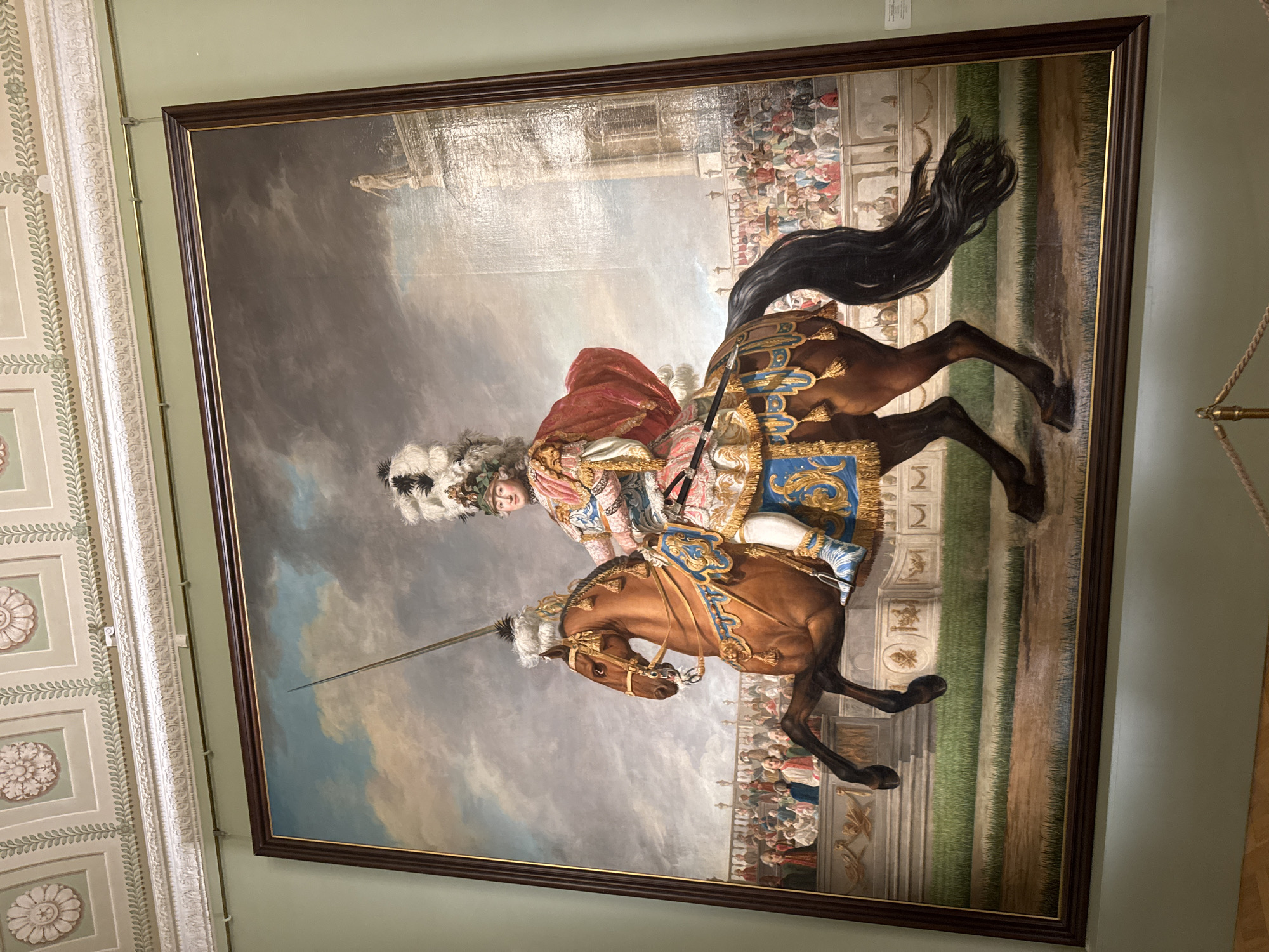 This is a full-length oil painting on canvas, depicting a man in elaborate ceremonial attire riding a brown horse. The rider wears a large feathered headdress, a richly decorated pink and blue costume with a red cape, and white boots. The horse is captured mid-stride, with its front left leg raised. Both the rider and the horse are adorned with ornate trappings.
In the background, a crowd of spectators is seated in tiered seating, suggesting a public event. Behind the spectators, there is a large, light-colored classical building. The sky above is dramatic with clouds.
The painting is housed in a dark wooden frame with gold trim. The artwork is displayed in a room with light green walls and a decorative ceiling adorned with floral motifs.
This painting is located in the **Hermitage Museum in Saint Petersburg, Russia**. The Hermitage Museum is one of the largest and oldest museums in the world, housed in a complex of buildings, including the Winter Palace. The painting's style suggests it is likely an equestrian portrait from the 18th century.
