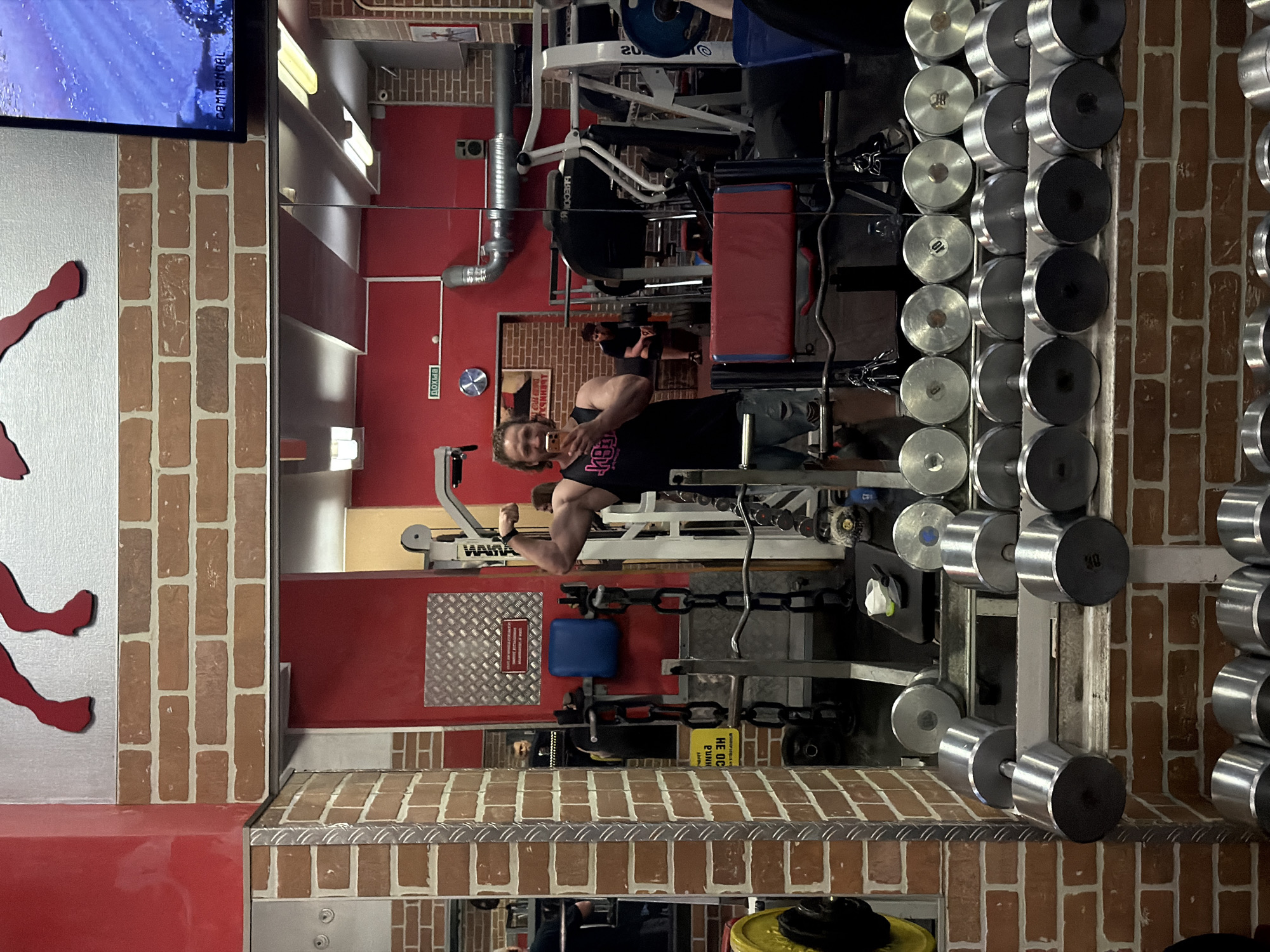The image depicts a man taking a mirror selfie in a gym. He is wearing a black tank top and ripped jeans, flexing his bicep. The gym environment features brick walls, metal equipment, and rows of dumbbells. In the background, other gym-goers are visible, some engaged in workouts and others resting. The gym appears to be located in Saint Petersburg, Russia, as suggested by the Cyrillic text visible on a sign in the background which reads "Выход" (Exit).