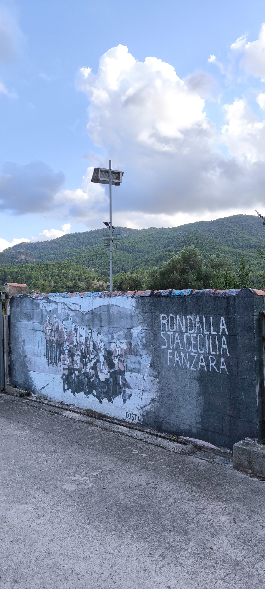 The image shows a mural on a weathered concrete wall in Fanzara, Spain. The mural depicts a group of people, likely musicians, holding instruments such as guitars. They are arranged in rows, with some standing and others seated. The style of the mural is somewhat faded and monochrome, with details of their clothing and poses visible. To the right of the mural, white text reads "RONDALLA STA. CECILIA FANZARA," indicating the subject of the artwork is likely a musical group named "Rondalla Santa Cecilia" from Fanzara. A signature "COSTI" is visible in black text at the bottom of the mural. The wall is situated outdoors, with a paved area in front of it. In the background, a landscape of green, forested hills rises under a partly cloudy sky. A tall metal pole with a solar panel and antennas is visible above the wall, against the sky. The overall scene suggests an outdoor public space in a rural or semi-rural area of Fanzara.