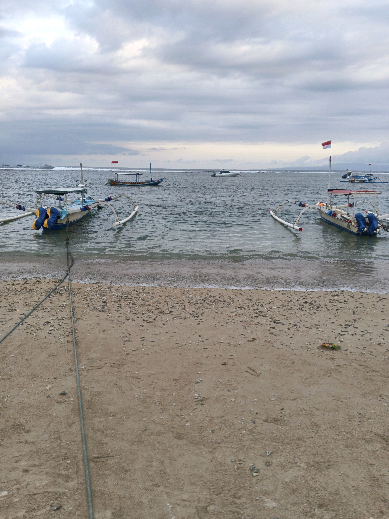 The image depicts a serene beach scene in Denpasar, Indonesia, under a cloudy sky. In the foreground, a sandy beach is visible, with a rope extending from the water and disappearing into the sand. Two traditional Indonesian fishing boats, known as jukung, are anchored in the shallow water. These boats are characterized by their outriggers, which provide stability. One jukung, on the left, has yellow and blue buoys attached and is secured by a rope to the shore. The other jukung, on the right, displays a red and white Indonesian flag on a pole. In the distance, several other boats, including a white speedboat and a larger vessel, are scattered across the calm sea. The overall atmosphere is peaceful, with the gentle waves lapping at the shore and the overcast sky creating a diffused light.