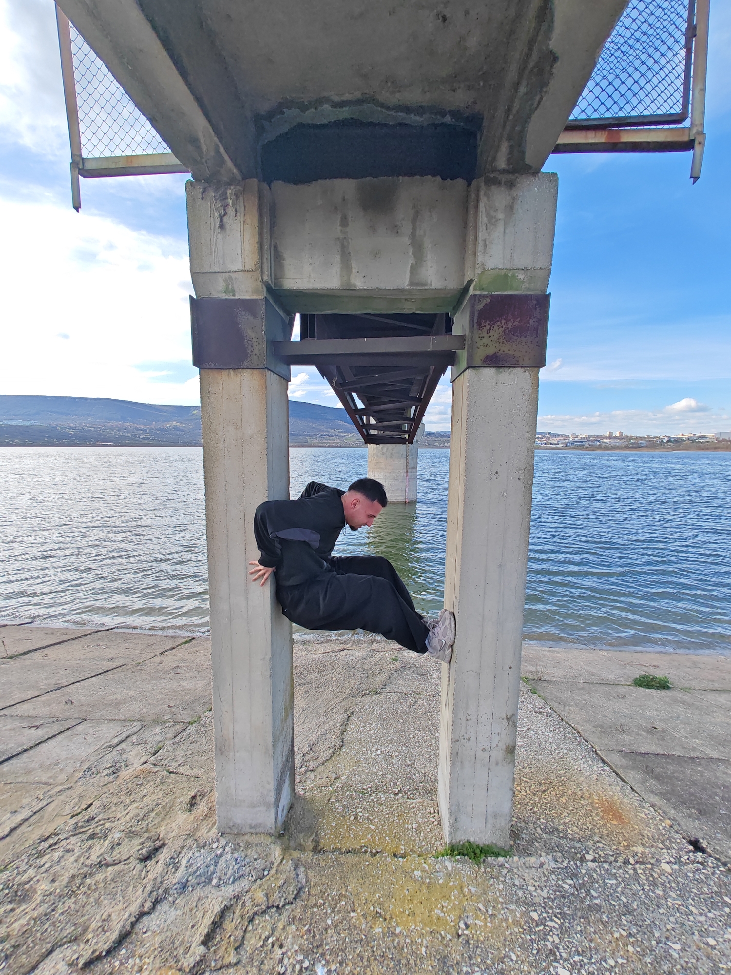 A man is seen performing a maneuver between two concrete pillars under what appears to be a bridge or overpass. The man is wearing dark clothing and light-colored sneakers. He's bent over, using his hands on one pillar and feet on the other to support himself. Beyond the structure, there is a body of water, likely a lake or reservoir, with land and buildings visible in the background. The sky is blue with scattered clouds. The scene is taking place near the city of Shumen, Bulgaria.
