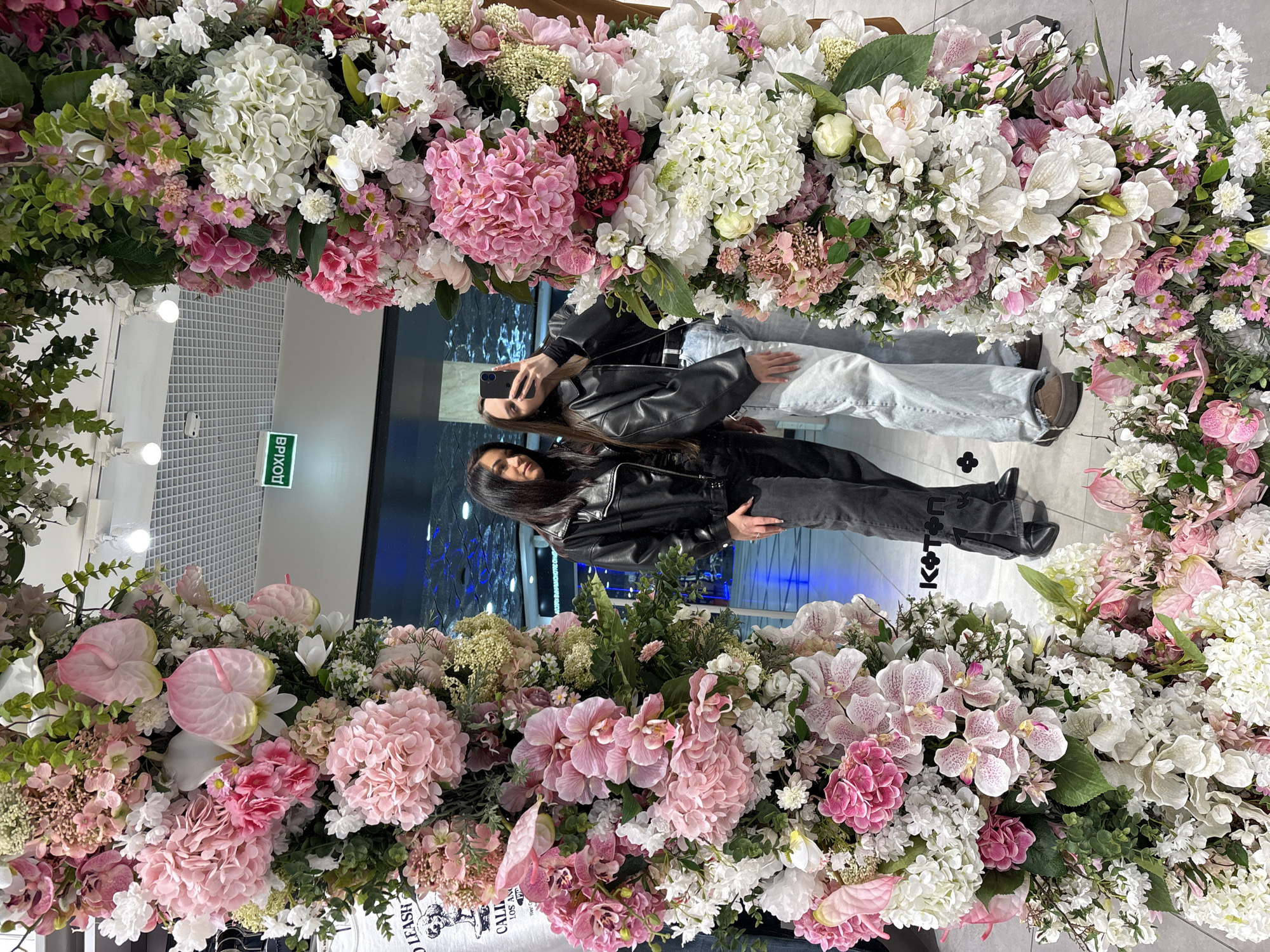 Two women pose for a selfie in front of a floral archway in Yekaterinburg, Russia. The woman on the left holds a phone and wears a black leather jacket and light blue jeans. The woman on the right also wears a black leather jacket and black jeans. The floral archway is composed of pink and white flowers. The scene is indoors, with a tiled ceiling and an emergency exit sign visible in the background.