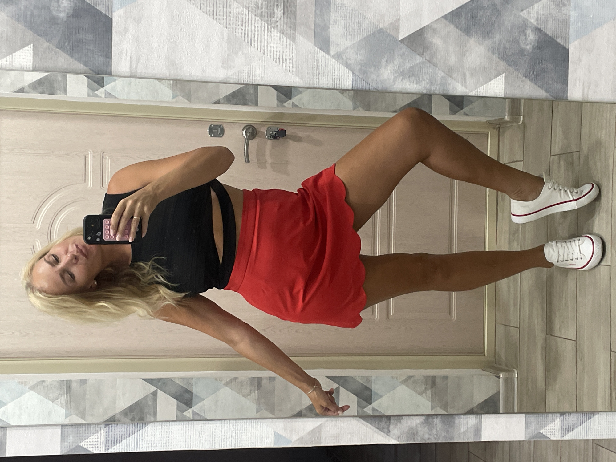 A blonde woman stands in front of a door and a mirror, taking a selfie. She is wearing a black crop top and a red scalloped skirt, with white sneakers. The background appears to be an interior hallway with tiled flooring and wallpaper. Based on the provided information, this scene is located in Novorossiysk, Russia.