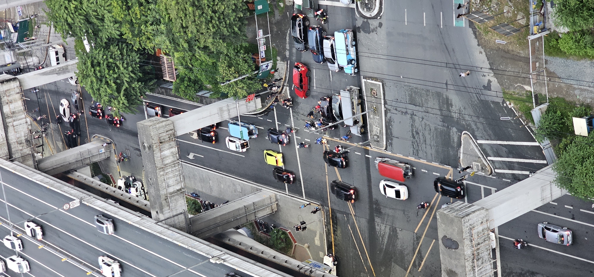 An aerial, vertical, full shot captures a busy urban intersection in Makati, Philippines, with layered overpasses and a dense urban environment. The scene is dominated by a multi-level elevated highway system, with traffic flowing on different levels. The lower level, where most of the activity is concentrated, is a wet asphalt road with yellow and white lane markings. Various vehicles are present, including cars, motorcycles, and a jeepney.
In the foreground, a red SUV is parked, and a black SUV is to its right. Several motorcycles are scattered throughout the intersection, some with riders. A light blue jeepney is also visible. Further back, on the lower level, a yellow car, a black sedan, and a white sedan are stopped at an intersection, possibly at a red light. Two individuals are standing near the intersection, one in a blue shirt and another in black.
On the level above, a white van is moving, and further up the road, a line of cars is visible on the elevated highway. The overpasses themselves are made of concrete, with some scaffolding or construction materials visible on the side of one. Lush green trees flank the right side of the image, suggesting a partially green urban landscape. The overall impression is one of a bustling, dynamic city environment with continuous movement and activity.