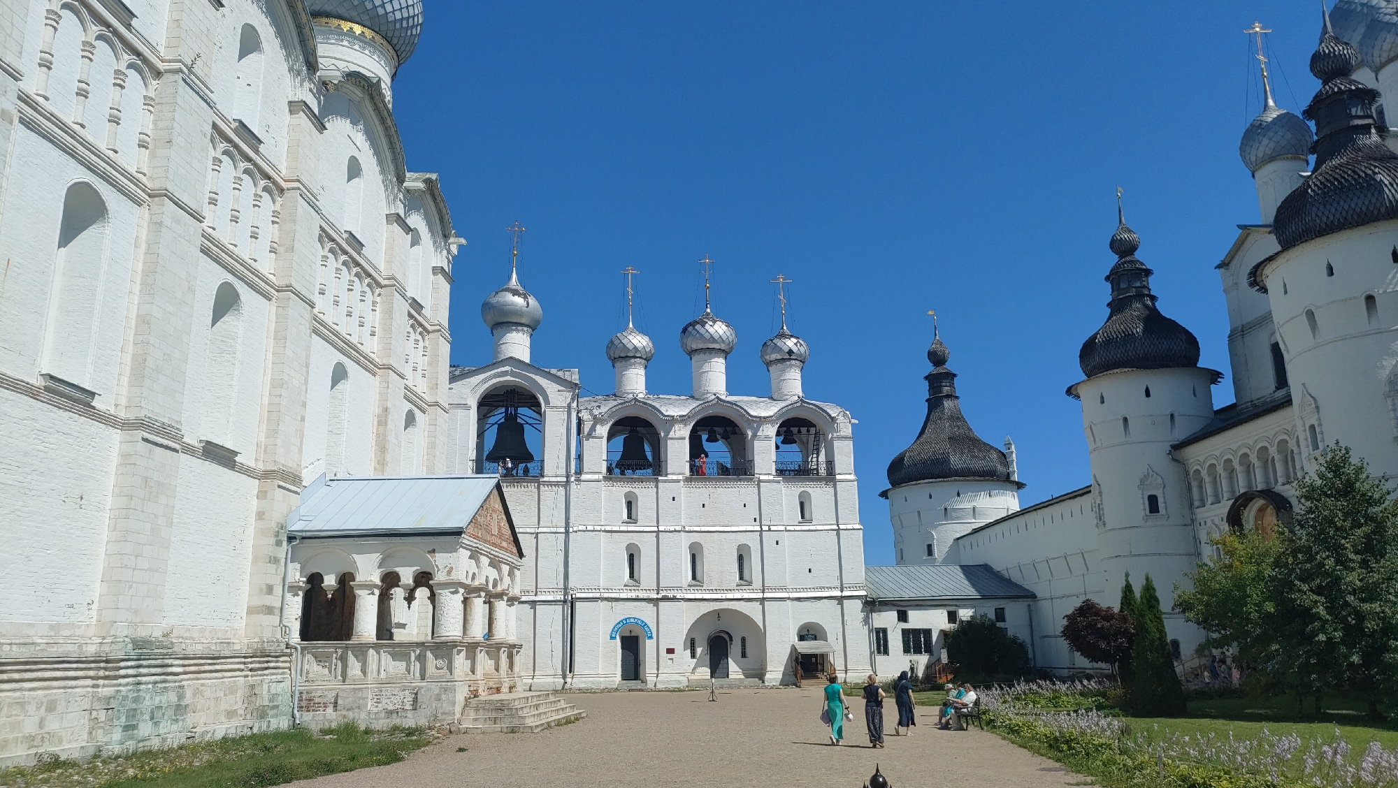 The image depicts the Rostov Kremlin in the city of Rostov, located in Yaroslavl Oblast, Russia. The scene is dominated by a large, white brick complex of religious buildings under a clear blue sky. In the foreground, a courtyard with a gravel surface is visible, with several people walking and sitting. To the left, a white, multi-story building with arched windows and decorative details is prominent. Adjacent to it is a smaller, single-story structure with a pitched roof.
The central focus is a tall bell tower with multiple arched openings, each containing a large bell. Above the bell tower are several onion domes topped with crosses. To the right of the bell tower, a larger building with a dark, conical roof and white, cylindrical towers rises. This building also features arched windows and decorative architectural elements.
Several individuals are present in the courtyard. Three women are walking towards the right side of the frame, one in a green dress, one in a dark blue dress, and another in a dark dress. Two people are seated on a bench in the lower right corner, facing away from the camera. Another person is standing near the bell tower, looking towards the bells. Lush green trees and flowering plants are visible in the middle ground and background, suggesting a well-maintained outdoor space. The architecture is characteristic of traditional Russian religious structures, with white walls, silver domes, and ornate detailing. The lighting suggests it is a bright, sunny day.