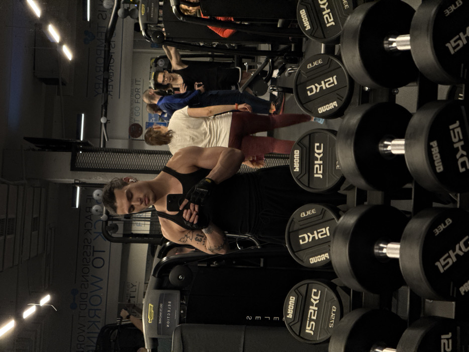 This image depicts an indoor gym environment, specifically a Pulse Fitness & Spa location in Sofia, Bulgaria, characterized by its equipment and aesthetic. In the immediate foreground, a multi-tiered dumbbell rack displays several black dumbbells, including weights labeled "12KG" and "15KG" (marked "PROUD"). Behind this, a rack holds black circular weight plates marked "J2KC" and "BEYOND," likely representing 20kg.
The central subject is a young adult male, positioned in the mid-foreground, appearing to take a mirror selfie. He has dark hair tied back, wears a black tank top, black track pants, and black fingerless gloves. A white earbud is visible in his right ear, and a tattoo is present on his left arm. He holds a black smartphone with both hands, pointing it towards his reflection.
The large mirror behind the subject reflects other gym patrons and equipment. A female in a white t-shirt and dark red leggings stands with her back to the camera. To her right, two males are visible: one with a bald or shaved head in a blue long-sleeved shirt, and another in a black tank top and glasses, holding a phone and leaning against a fitness machine. A partial view of a person in a red top is visible on the far right. Technogym brand exercise machines are present in the background. Overhead strip lights illuminate the space and are reflected in the mirror, along with reversed text on the wall or mirror, including phrases like "BACK TO WORKING" and "IT'S TEMPORARY." The gym features a dark color palette with industrial elements.