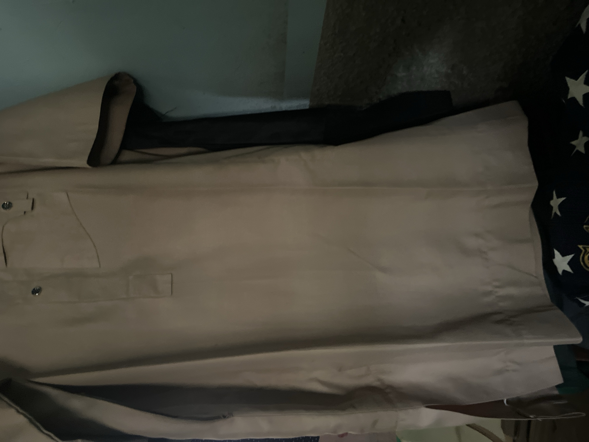 The image shows a close-up, overhead view of a beige garment, possibly a kaftan or a similar traditional tunic. The fabric appears to be smooth and draped, suggesting it's made of a lightweight material. The garment is laid out flat, with some folds and creases visible, indicating it's either being prepared for wear or has been recently taken off.
On the left side of the garment, there are two buttons and a pocket. The pocket flap is slightly open, revealing a darker interior. A rectangular tab is also visible on the chest area, possibly for a decorative element or to secure the garment. The background is a dark, textured surface, possibly a wall or a floor, with a hint of a teal-colored surface on the far left, possibly another wall or fabric. To the right of the beige garment, a dark fabric with white star patterns is partially visible, suggesting another item of clothing or bedding.
There are no people visible in the image, nor any clear indication of time of day, weather, or specific emotions. The setting appears to be indoors, given the lighting and the draped fabric. The image is likely taken in Jauro Yinu, Nigeria, as per the location context provided, which might imply the garment is of local origin or style. The overall impression is one of stillness and focus on the texture and details of the garment itself.