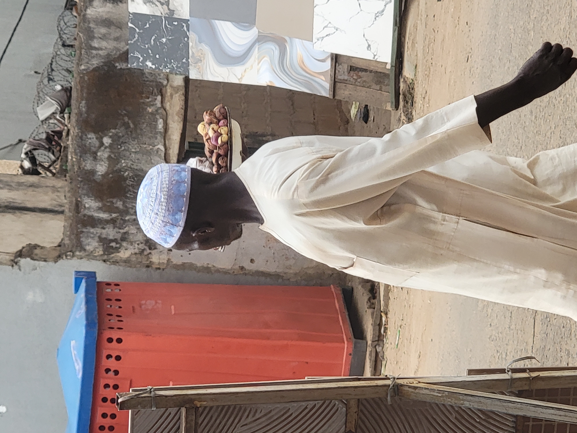 An elderly Black man wearing a white robe and a blue patterned skullcap walks on the side of a road in Maiburiji, Nigeria. On his head, he carries a white dish piled with small red onions. Behind him are orange and blue plastic containers. Atop those containers is a stack of square tiles with swirling marbleized patterns. A low, weathered, wooden structure with corrugated tin is visible in the foreground, and there is a cement wall behind the man and containers. The environment suggests an outdoor marketplace.
