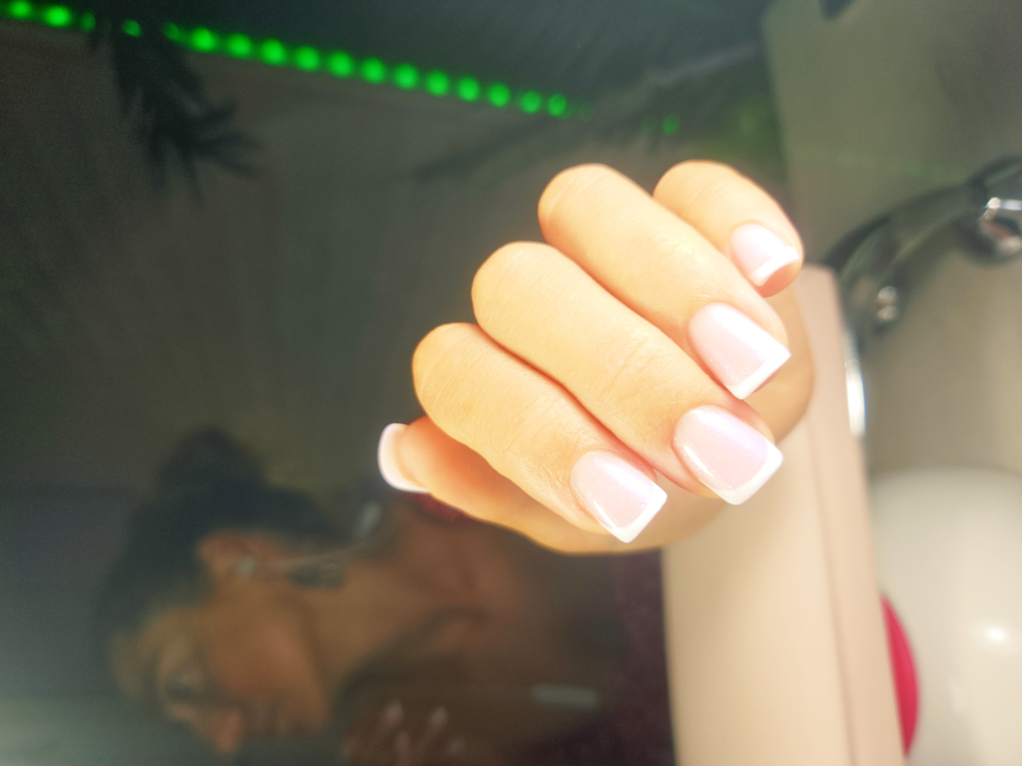 A close-up, over-the-shoulder shot shows a woman's hand with neatly manicured French tip nails. The woman is out of focus in the background, wearing earrings and looking to the left. To the right of the hand, a string of green LED lights is visible, casting a glow. In the foreground, a portion of a pink and white nail lamp is visible. The scene appears to be indoors.
