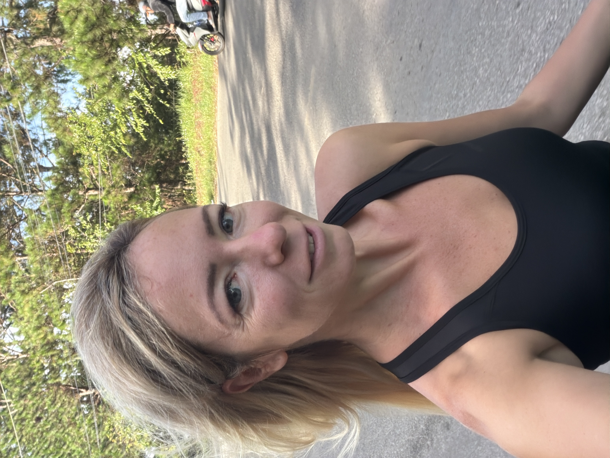 In this selfie taken outdoors, a blonde woman with fair skin is looking at the camera and smiling. She is wearing a black tank top. Her hair is blowing to the left, partially obscuring her face. In the background, there is a road and a grassy area with trees and foliage. A person on a scooter is visible in the distance on the right side of the road, traveling away from the camera. The scene appears to be on a road in a rural or suburban area. The location is identified as Xuân Trường - Đà Lạt, Vietnam.