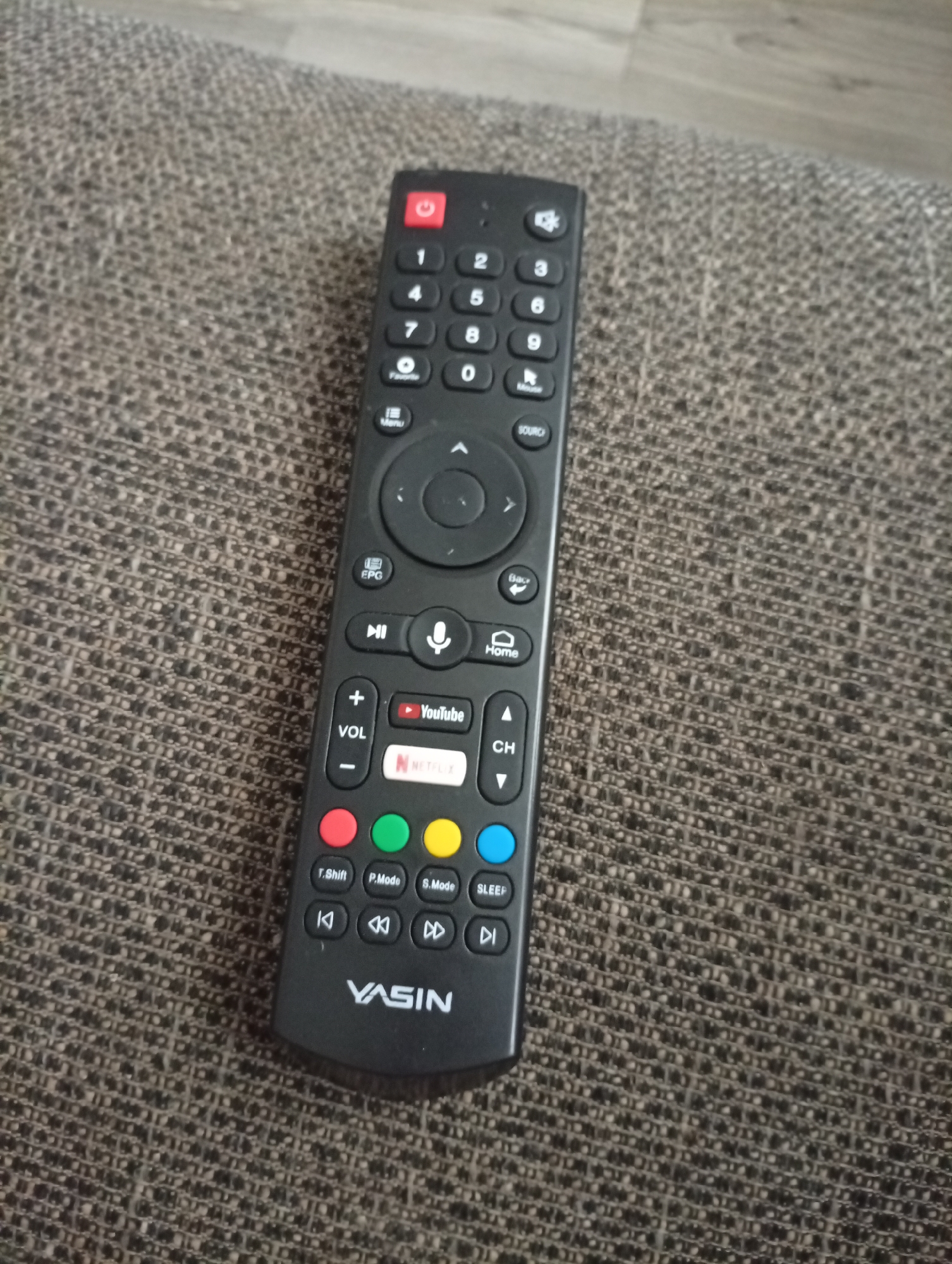 A black YASIN brand TV remote control is lying on a textured gray fabric surface. The remote has a power button at the top, followed by a numeric keypad (0-9), and directional buttons surrounding a central selection button. Below the numeric keypad are buttons labeled "Favorite", "Menu", and "Source". Further down are buttons for "EPG", "Mute", a microphone icon, and "Home". There are also volume up and down buttons, and buttons for YouTube and Netflix. The lower section of the remote has colored buttons (red, green, yellow, blue) and playback controls. The image is taken from a slightly elevated angle, focusing on the remote. The background shows a blurred gray and brown textured surface, likely upholstery. The location of Semetey, Kyrgyzstan is not discernible from the image itself.