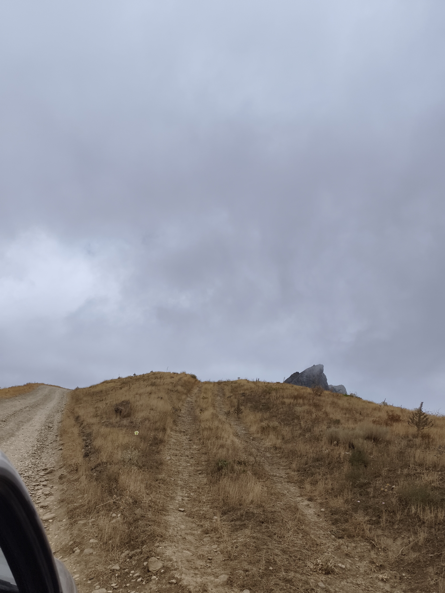 The image depicts a dirt road winding up a dry, grassy hill under a cloudy sky. In the distance, a rocky peak emerges from a light mist. The scene appears to be in Qalaşıxı, Azerbaijan, a region known for its mountainous terrain and historical sites. The road is rutted, suggesting it is used by vehicles. The vegetation is predominantly dry grass and sparse bushes, indicating an arid or late-season environment. The overall atmosphere is muted and overcast. No people or specific man-made structures are visible, other than the road itself.