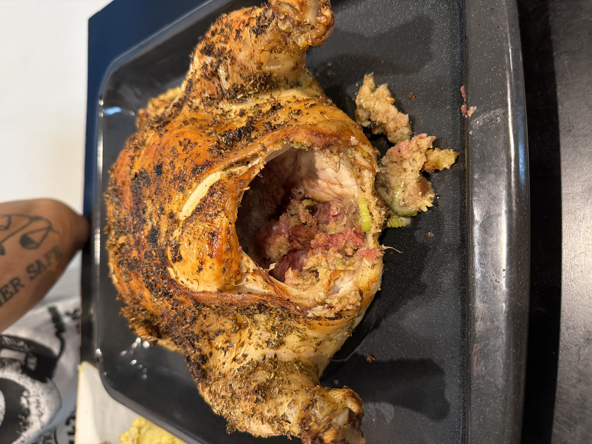 A roasted chicken, stuffed with stuffing, is on a black baking pan. The chicken's skin is browned and seasoned. The stuffing appears to contain celery and ham. An arm with a tattoo reading "Never Safe" is partially visible on the left side of the frame. The background appears to be a kitchen countertop. No evidence suggests that this is in Los Angeles.