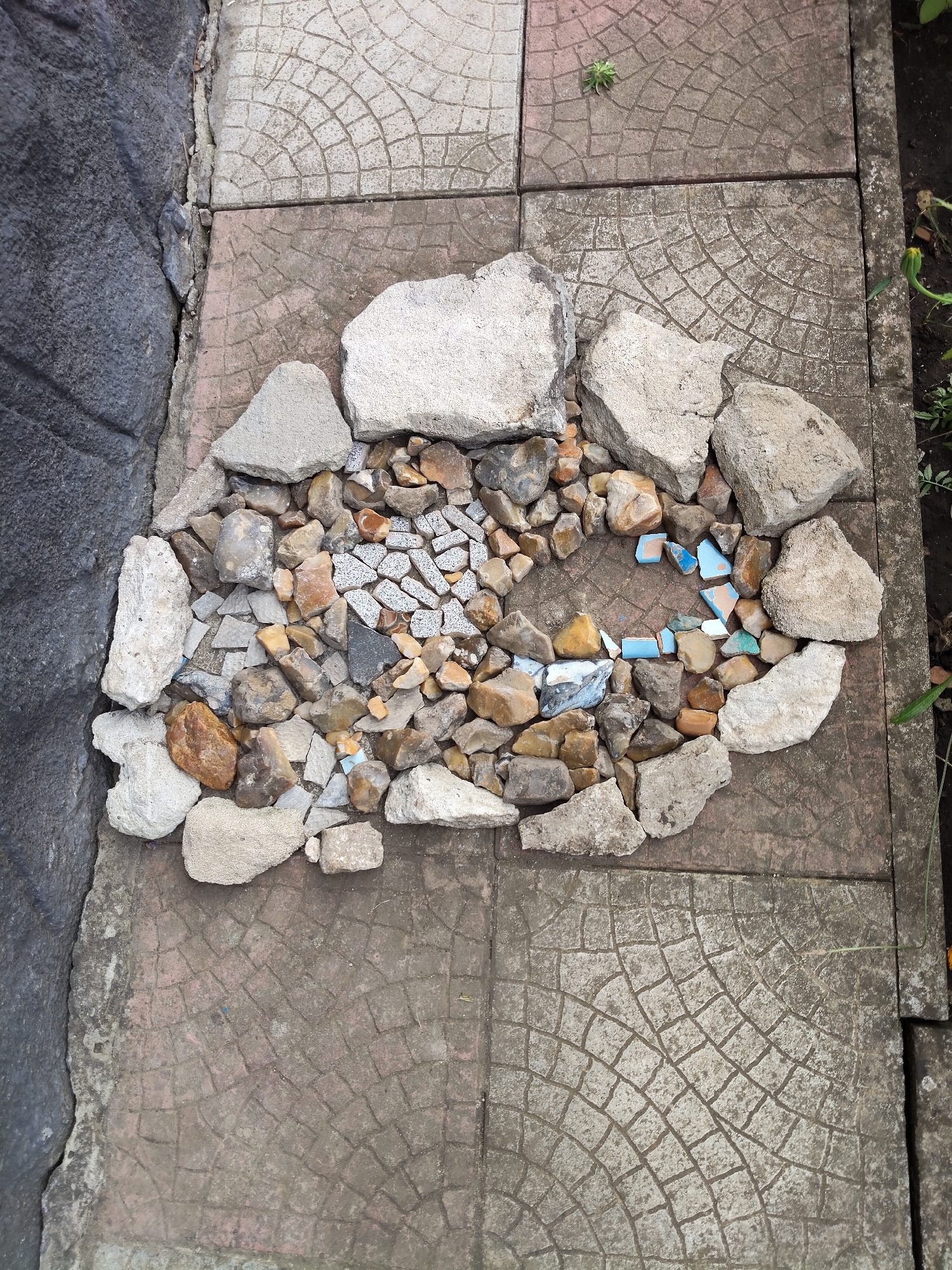 A cluster of rocks and broken tiles forms a mosaic-like arrangement on patterned paving stones. The paving stones are laid out in a grid with a fan-like pattern etched into their surface. To the left, a dark, rough-textured wall meets the paving stones. A few small green plants are visible to the right and in the upper right corner of the frame, suggesting an outdoor setting. The overall scene depicts an outdoor area with an artistic arrangement of natural and man-made materials. This location is Gîrbova, Moldova.