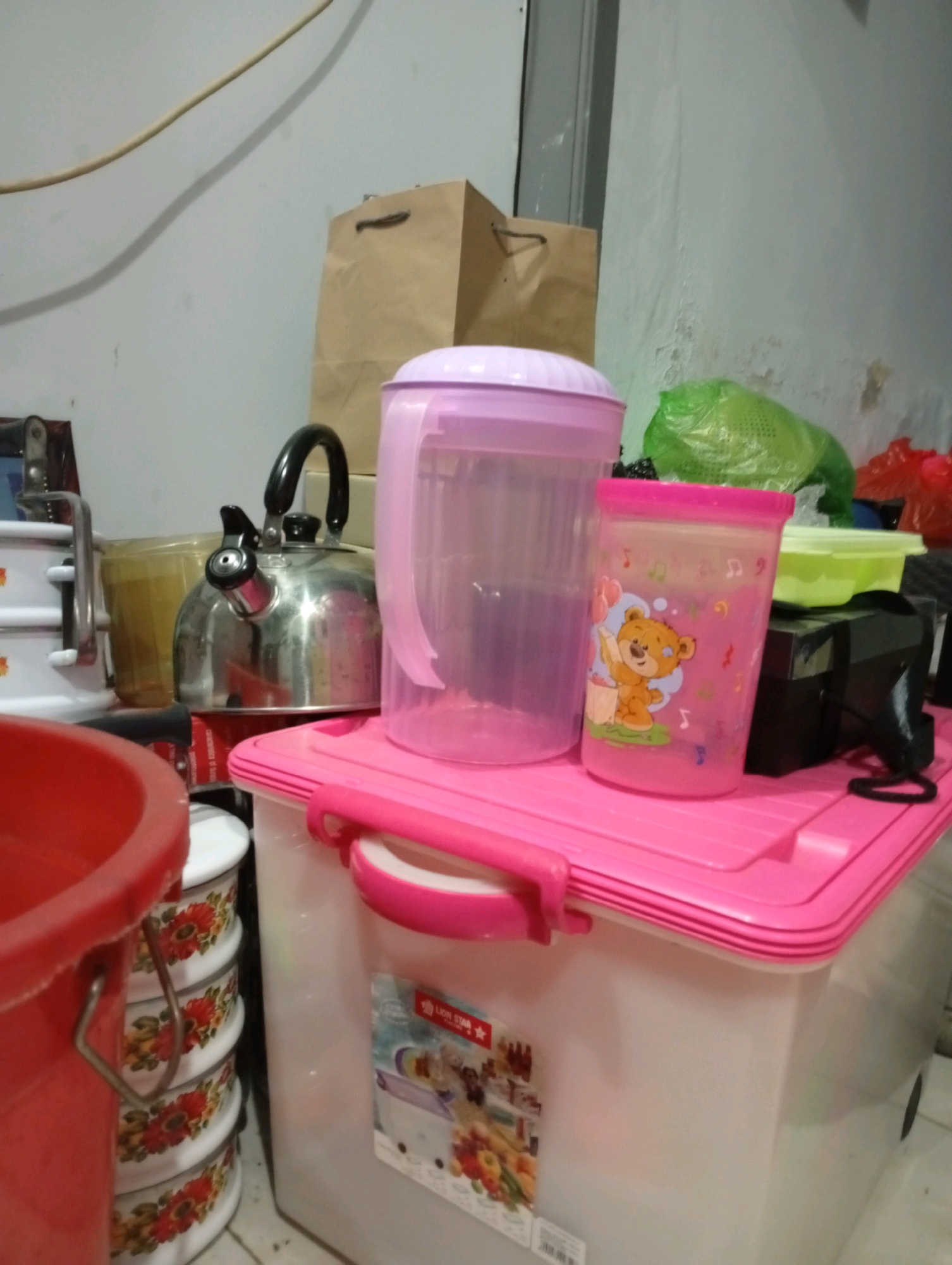 The image shows a collection of household items on a table, likely in a storage area or a kitchen in Kebondalem, Indonesia. In the foreground, a large red bucket is partially visible on the left. To its right, a stack of cylindrical containers with floral patterns is neatly arranged. Behind these, a stainless steel kettle with a black handle sits on what appears to be a red box.
Dominating the center of the image is a large, translucent pink plastic jug with a purple lid. Next to it, a smaller pink plastic cup with cartoon characters, including a bear, is also visible. Further back, a green plastic bag, possibly containing vegetables, and a red plastic bag are present. A brown paper bag stands upright in the background. The overall impression is of a domestic environment with various storage and kitchenware items.