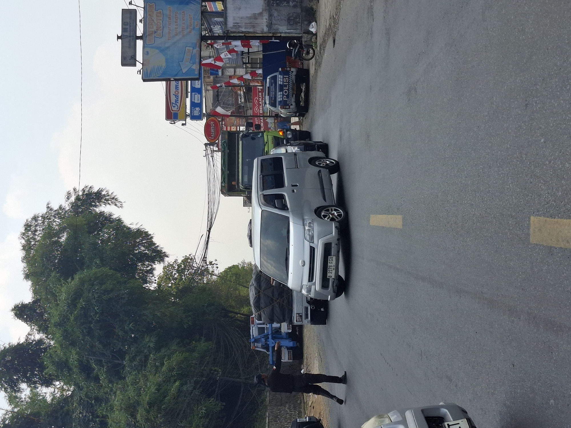 A traffic officer in a black uniform and cap directs traffic on a two-lane asphalt road in Pamuruyan, Indonesia. The officer faces left with their right arm extended, gesturing for vehicles to stop or proceed. In the foreground, a silver minivan with license plate "B 2178 FGV" is stopped, facing forward. Behind the minivan, a light truck with a green cab and a trailer loaded with a large black tarp is stopped. Further back, a blue tow truck is visible. On the right side of the road, a white police vehicle with "POLISI" written on its side is parked. Several commercial signs are visible in the background, including "Indomart" and a sign for "WATERPARK & HOTEL." The scene is set against a backdrop of lush green trees and a cloudy sky.