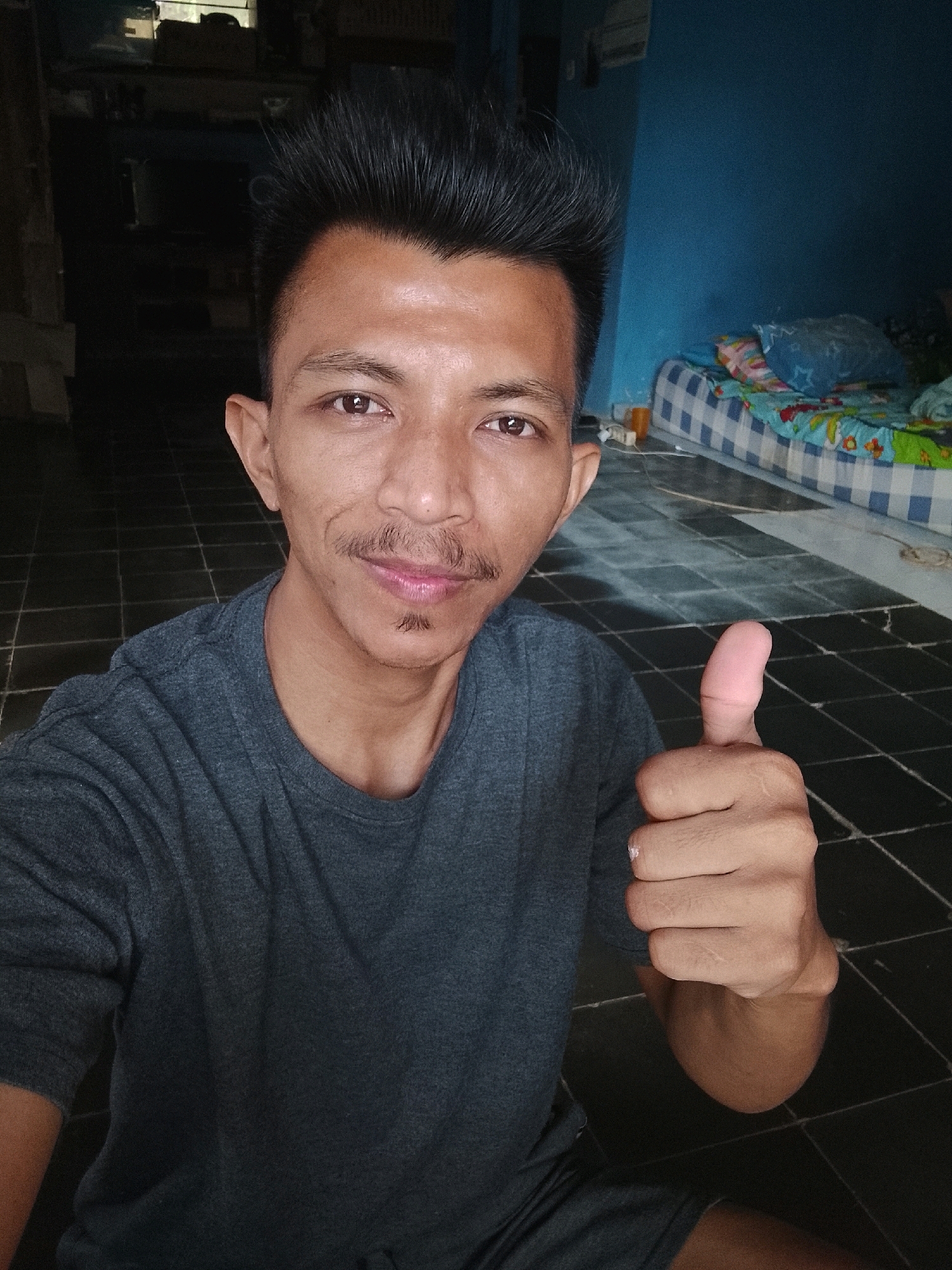 In Kertahayu, Indonesia, a young man with dark, styled hair and a mustache is pictured from the chest up, looking directly at the camera. He is wearing a dark gray t-shirt. His right hand is raised, with his thumb extended upwards in a "thumbs-up" gesture. He is positioned in what appears to be a room with a tiled floor, possibly a bedroom, as a bed with blue and white checkered bedding and colorful pillows is visible in the background. The walls are painted a vibrant blue. The lighting suggests an indoor setting, possibly during the day or evening.