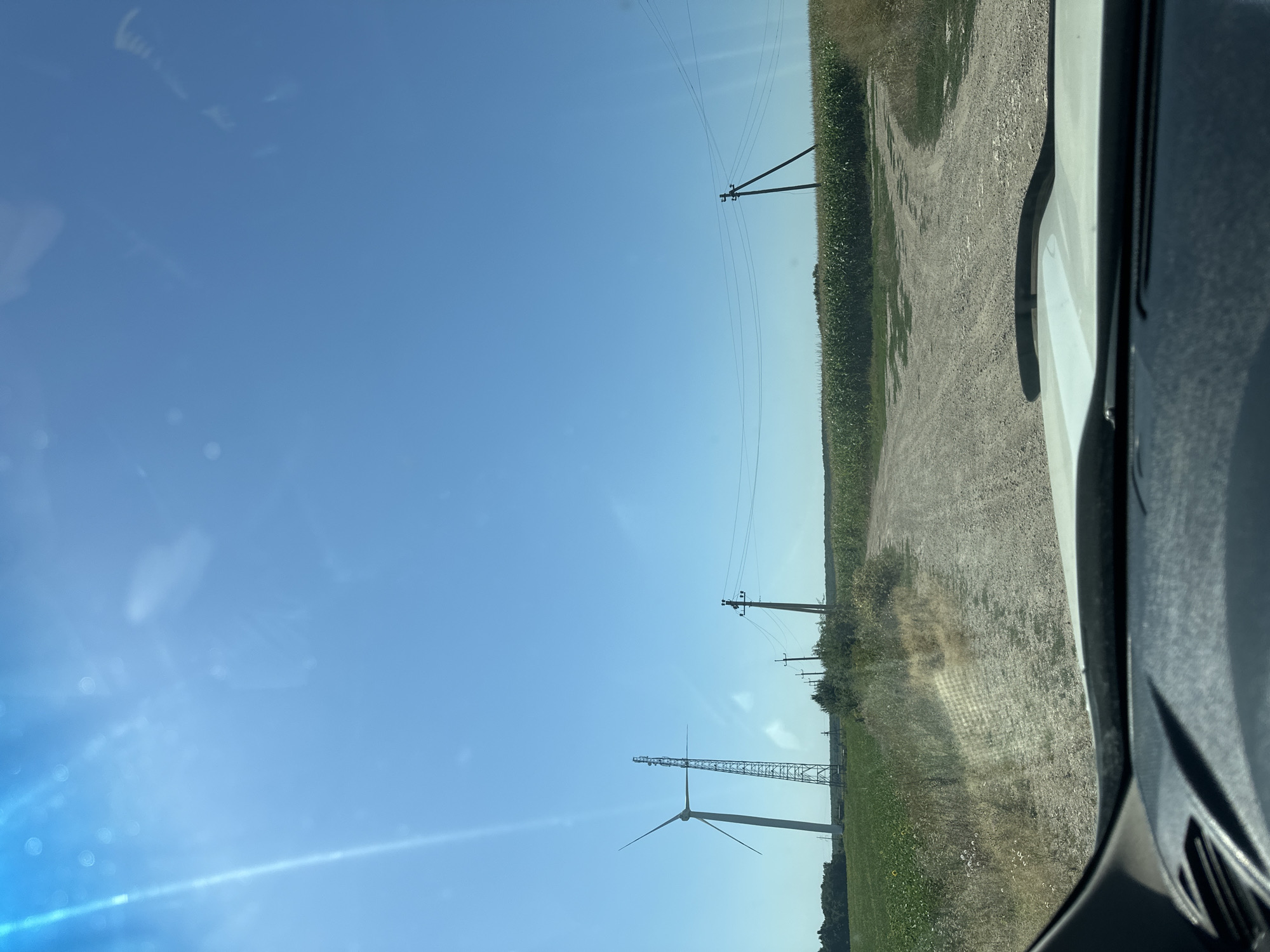The image depicts a rural scene on a clear, sunny day, likely in the city of Korchivka, Ukraine, viewed from the perspective of someone inside a vehicle. A dirt road forks ahead, leading into a landscape dotted with agricultural fields and utility infrastructure. To the left, a tall wind turbine stands prominently in a green field. Nearby, a metal tower with antennae is visible. Further to the right, a series of utility poles with wires stretch across the horizon, marking the path of power lines. The fields on either side appear to be crops, with one side a lighter green, possibly soybeans or a similar plant, and the other side a darker green, likely corn. The road surface is gravelly and uneven, with dry grasses and weeds growing along its edges. The sky is a bright, cloudless blue, with a visible lens flare originating from the upper left quadrant, indicating the direction of the sun. The foreground shows the dashboard and windshield of a vehicle, suggesting the image was captured while driving.