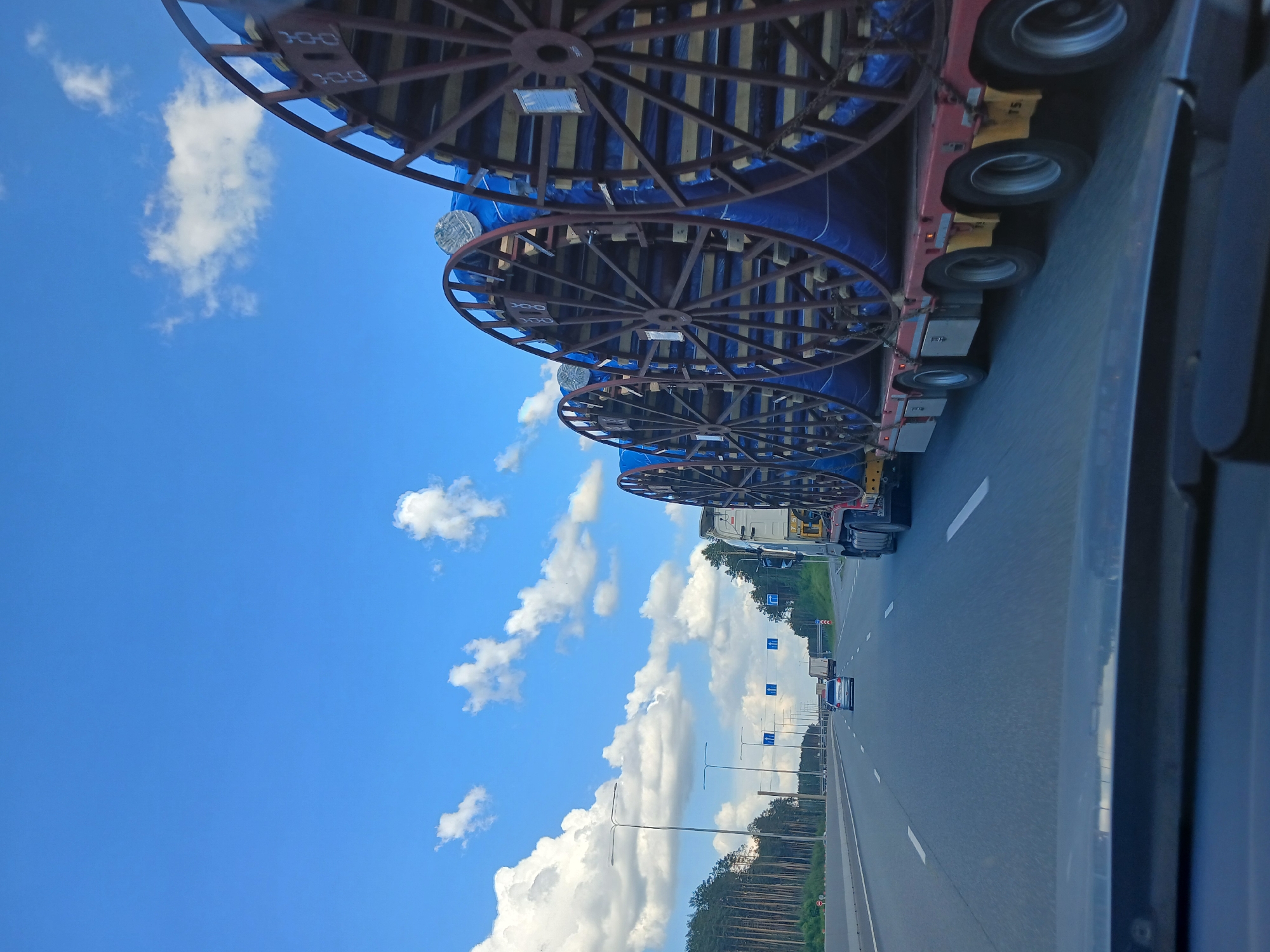 The image depicts a truck hauling several large spools, likely containing cable, on a highway. The spools are covered in blue tarpaulin and secured to a flatbed trailer. The truck is in the right lane of a multi-lane highway, with other vehicles visible in the distance. The highway is bordered by a dense forest on the left and a clear blue sky with scattered clouds on the right. The scene appears to be captured from a vehicle in motion, possibly from the perspective of a passenger. The image does not contain enough information to determine the exact location within Yekaterinburg, Russia, or specific details about individuals.