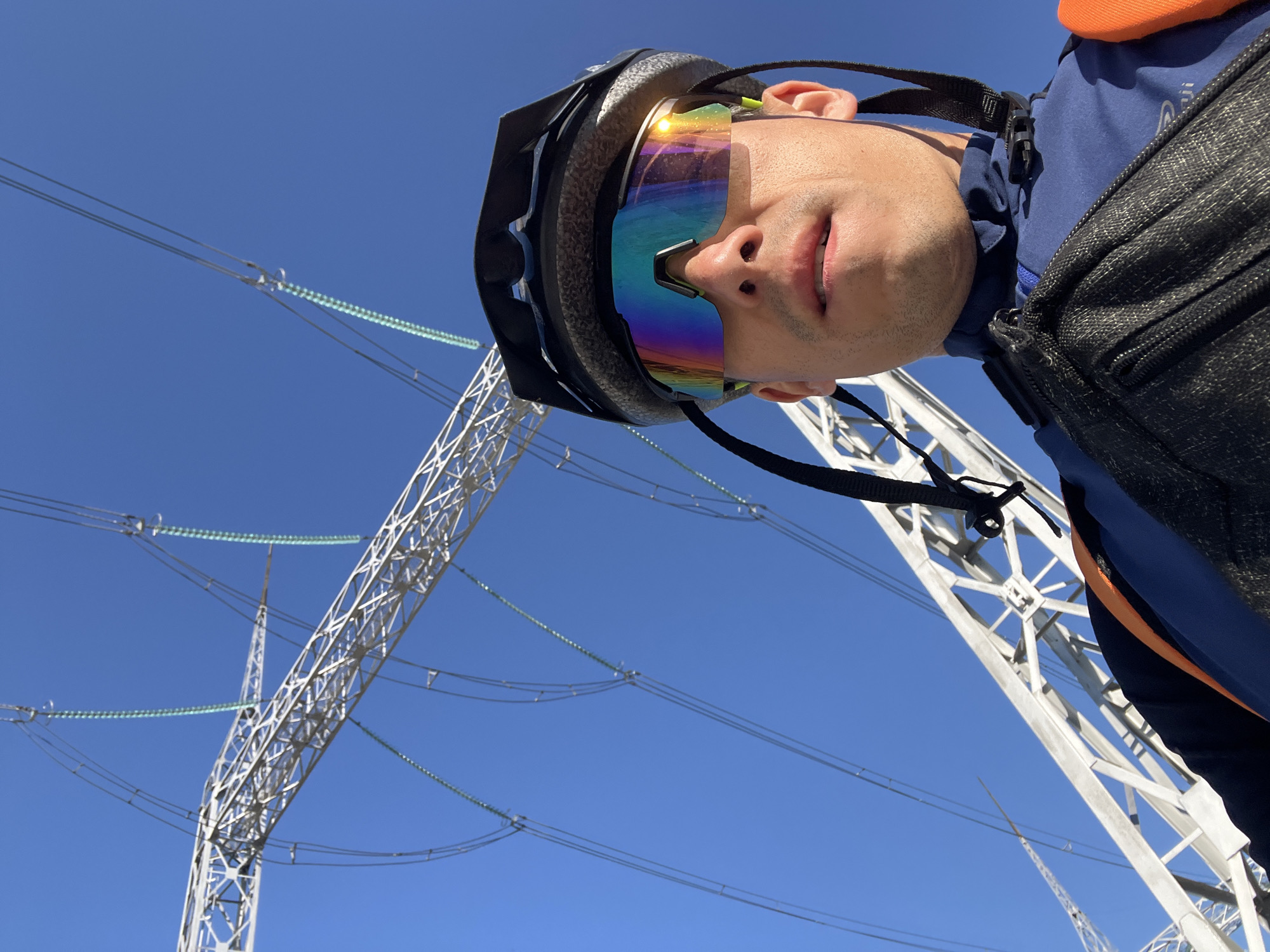 A man wearing a bicycle helmet and mirrored sunglasses takes a selfie outdoors under a bright blue sky. He is wearing a blue long-sleeved jacket over an orange safety vest and a black fanny pack across his chest. In the background, large metal power line towers and thick electrical cables are visible. The image appears to be taken at a low angle, looking up at the man and the power lines. The specific location in Chaikovsky, Russia, is not identifiable from the visual information alone, but the presence of extensive electrical infrastructure suggests an industrial or utility area.