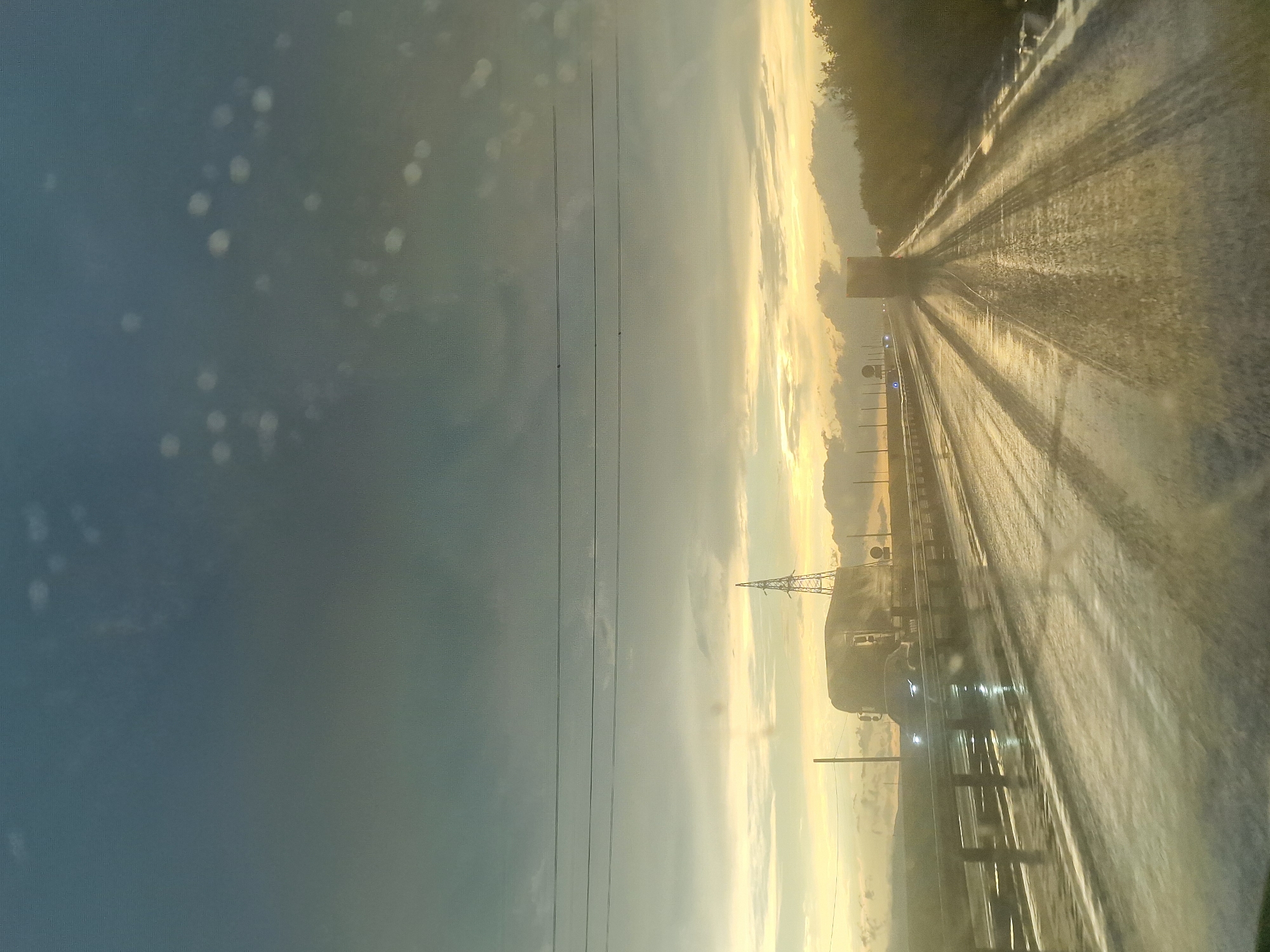 The image depicts a highway in Kursavka, Russia, under a stormy sky at dusk. The road is wet and reflective, with tire tracks visible from previous vehicles. The lighting suggests a low sun position, casting long shadows and creating a dramatic atmosphere. A truck with headlights illuminated is driving on the road. In the distance, a power transmission tower stands tall against the horizon. The overall scene conveys a sense of travel during challenging weather conditions.