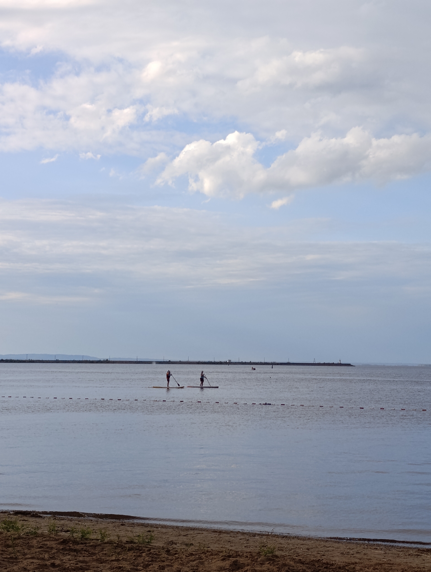 This image depicts a serene outdoor scene on a body of water, likely a lake or calm sea, under a cloudy sky. The foreground features a sandy beach with sparse vegetation. The main subjects are two individuals on paddleboards, actively engaged in stand-up paddleboarding. One person is wearing a bright orange life vest and the other a darker top. They are positioned some distance apart in the water.
In the background, a long, man-made structure, possibly a breakwater or pier, stretches across the horizon. Visible on this structure are several posts, and at the far right end, a lighthouse or navigational beacon. The water is calm, reflecting the overcast sky. A line of buoys or markers is visible in the water between the shore and the paddleboarders.
The time of day appears to be late afternoon or early evening, given the soft, diffused light. The weather seems mild, with scattered clouds in the sky, suggesting it's not actively raining but not a completely clear day. The overall mood is peaceful and recreational. There is no visible text in the image.