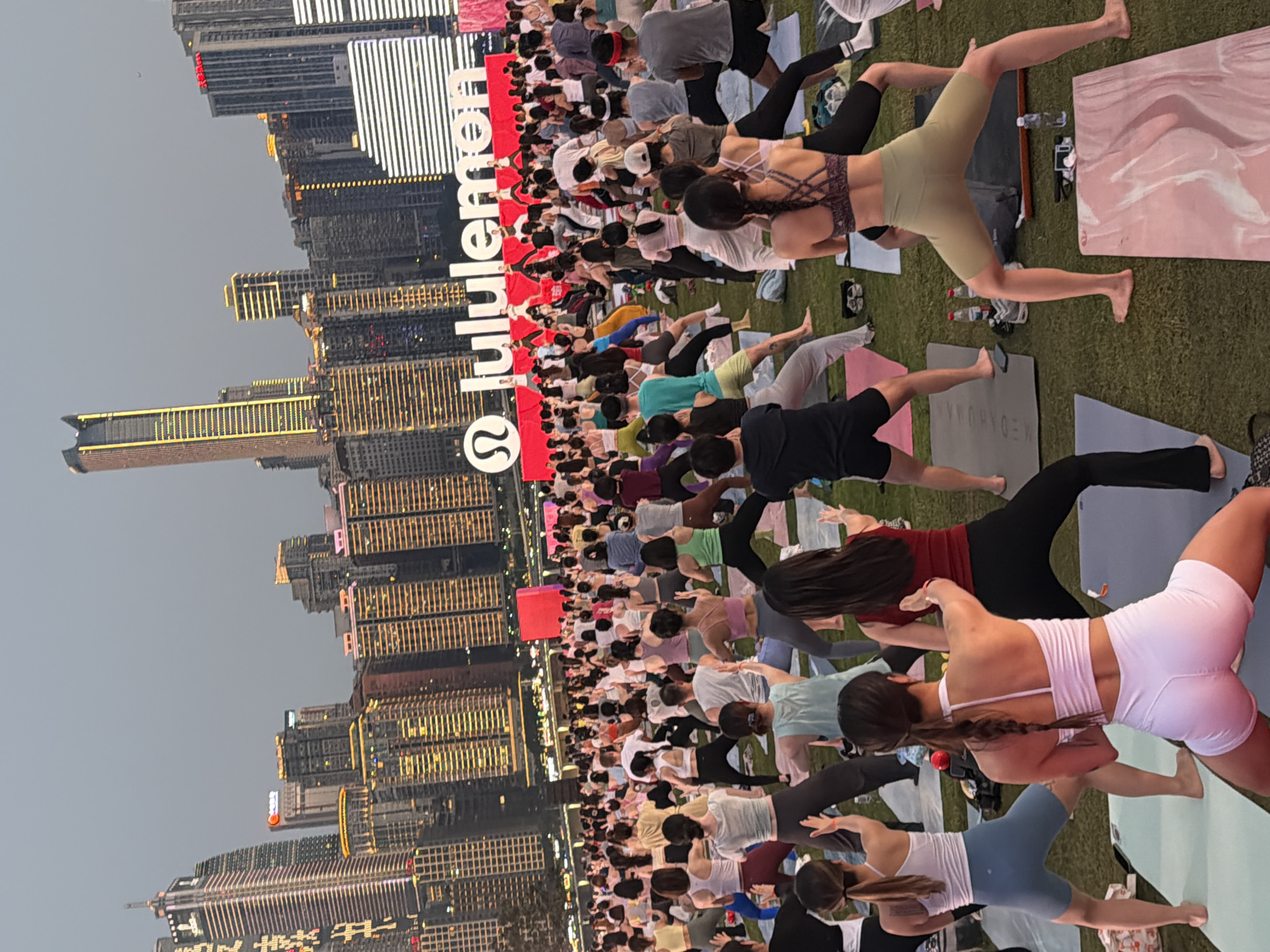 An outdoor yoga class is in progress on a grassy area in front of a city skyline at dusk. A large crowd of people, mostly women, are participating in the yoga session, performing various poses on yoga mats. In the background, a massive Lululemon logo is prominently displayed on a white structure, with the brand's name in large lettering. Behind the logo, a dense urban landscape of modern skyscrapers with illuminated windows stretches across the horizon. The sky is overcast, indicating either early evening or a cloudy day. The scene appears to be a promotional event or a large-scale community yoga gathering. The location is identified as Jiangbeicheng Street, China.