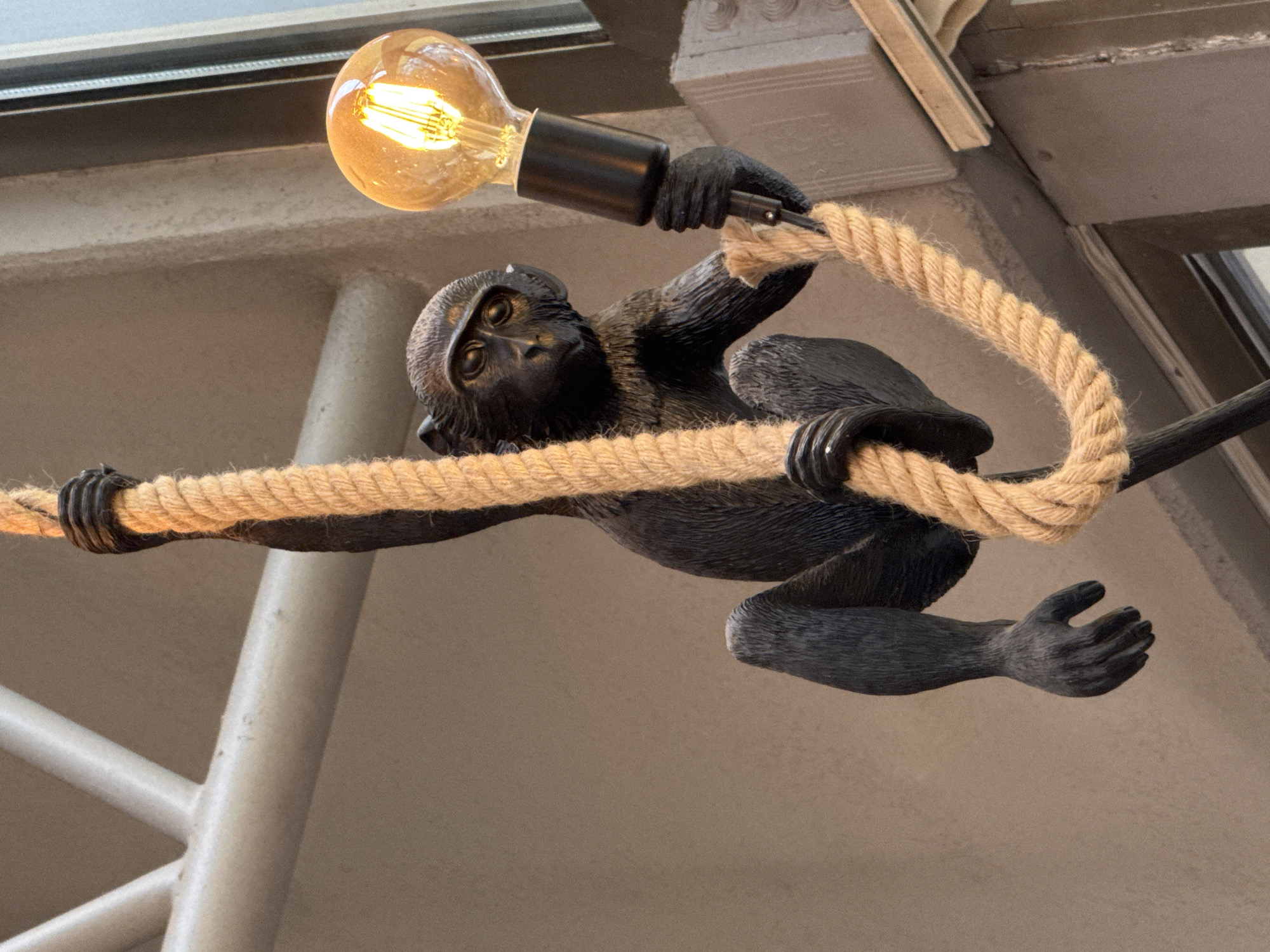 An indoor shot captures a creative lighting fixture featuring a dark monkey sculpture. The monkey, appearing to be made of resin or a similar material with a textured, fur-like finish, is depicted in a dynamic, playful pose, as if swinging. Its left hand and left foot are wrapped around a thick, braided beige rope that spans horizontally across the frame. With its right hand, the monkey holds the black socket of a glowing Edison-style lightbulb, which hangs above its head. The lightbulb emits a warm, bright yellow-orange light from its visible filament. The monkey's face is turned slightly towards the viewer, revealing dark, reflective eyes.
The background consists of muted, neutral tones of grey and beige, suggesting architectural elements like walls or ceilings, with some metallic or light-colored structures visible, including a possible window frame or ceiling beam in the upper left and a vertical support in the lower left. The overall lighting is warm, emanating primarily from the lightbulb itself, creating a cozy or atmospheric ambiance. No discernible text is visible in the image. The scene is located in Bishkek City, Kyrgyzstan.