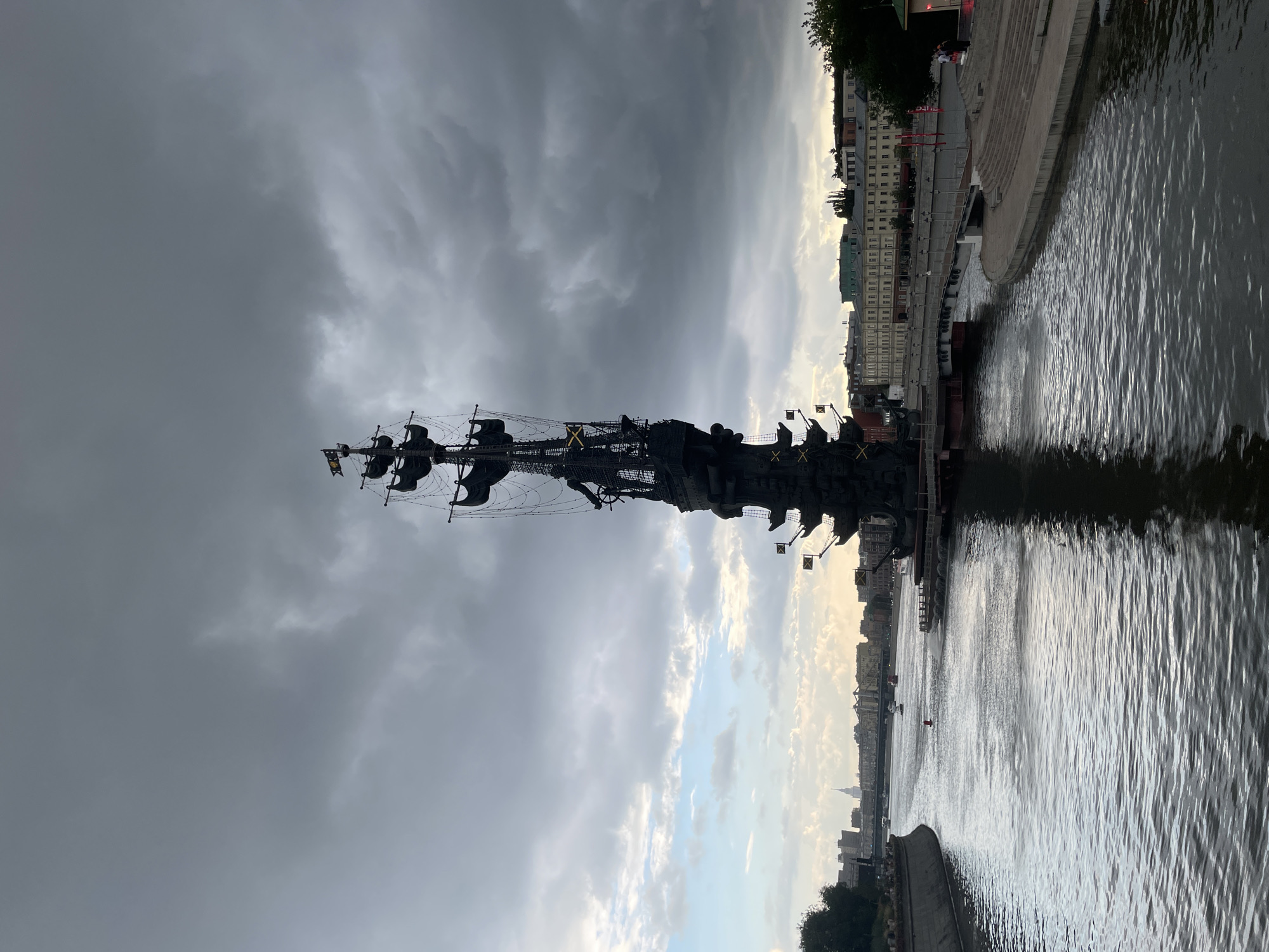This image depicts the Peter the Great Statue in Moscow, Russia. The colossal bronze monument, designed by Zurab Tsereteli, stands on an artificial island in the Moskva River, commemorating the 300th anniversary of the Russian Navy. The statue, which is over 300 feet tall, features Peter the Great standing atop a ship's hull, surrounded by sails and naval motifs. The statue is positioned in the foreground, with the river flowing in front of it and a city skyline visible in the background under a cloudy sky. The scene is illuminated by natural light, suggesting it was taken during the daytime.