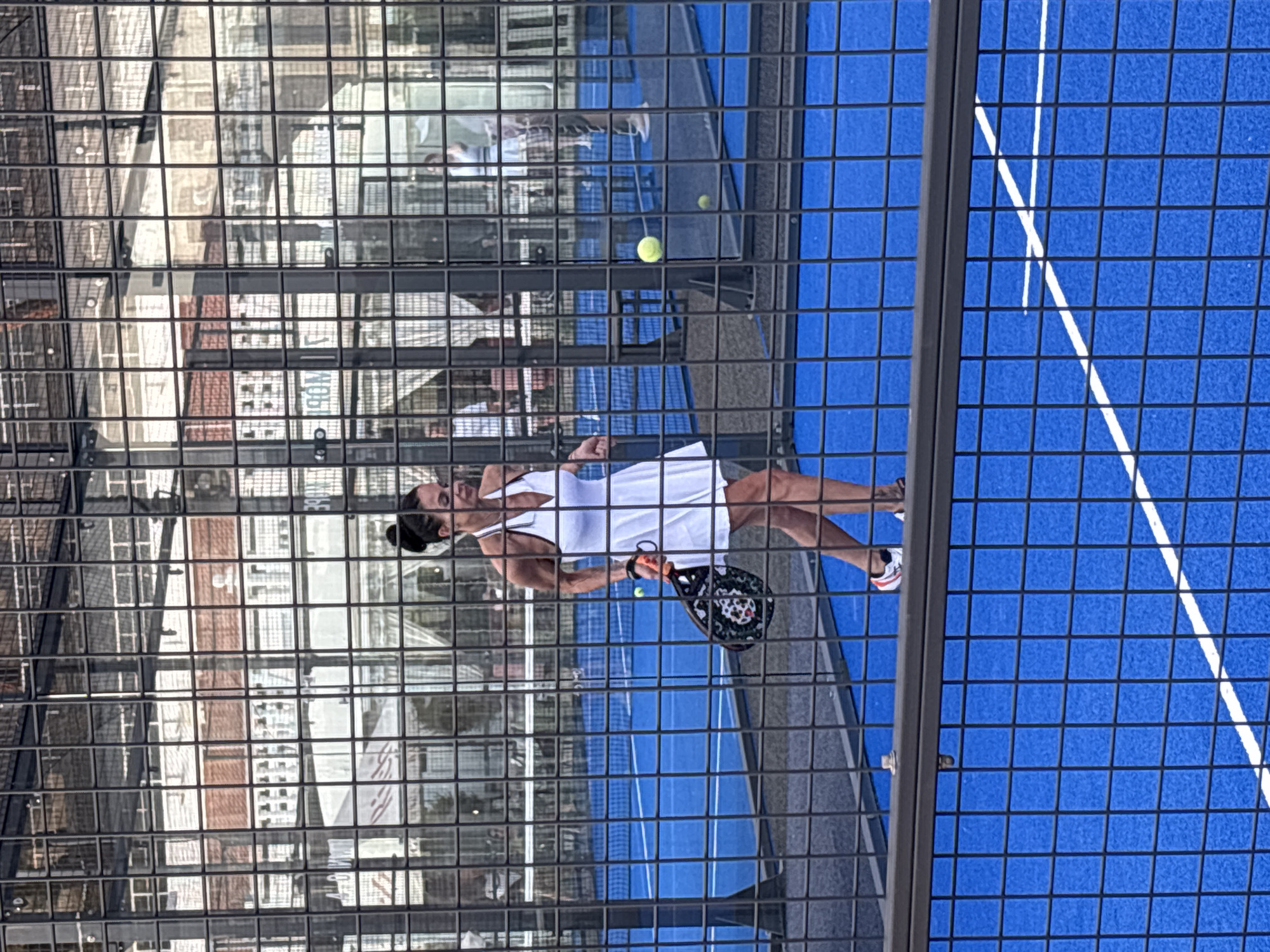 A woman in a white tennis dress with a halter neckline is playing padel on a blue court. She is holding a black and white padel racket. Two yellow padel balls are visible on the court. The court is enclosed by a metal fence. In the background, there are other players and buildings. The scene appears to be an outdoor sports facility. The specific location in the city of Sochi, Russia, is not discernible from the image.