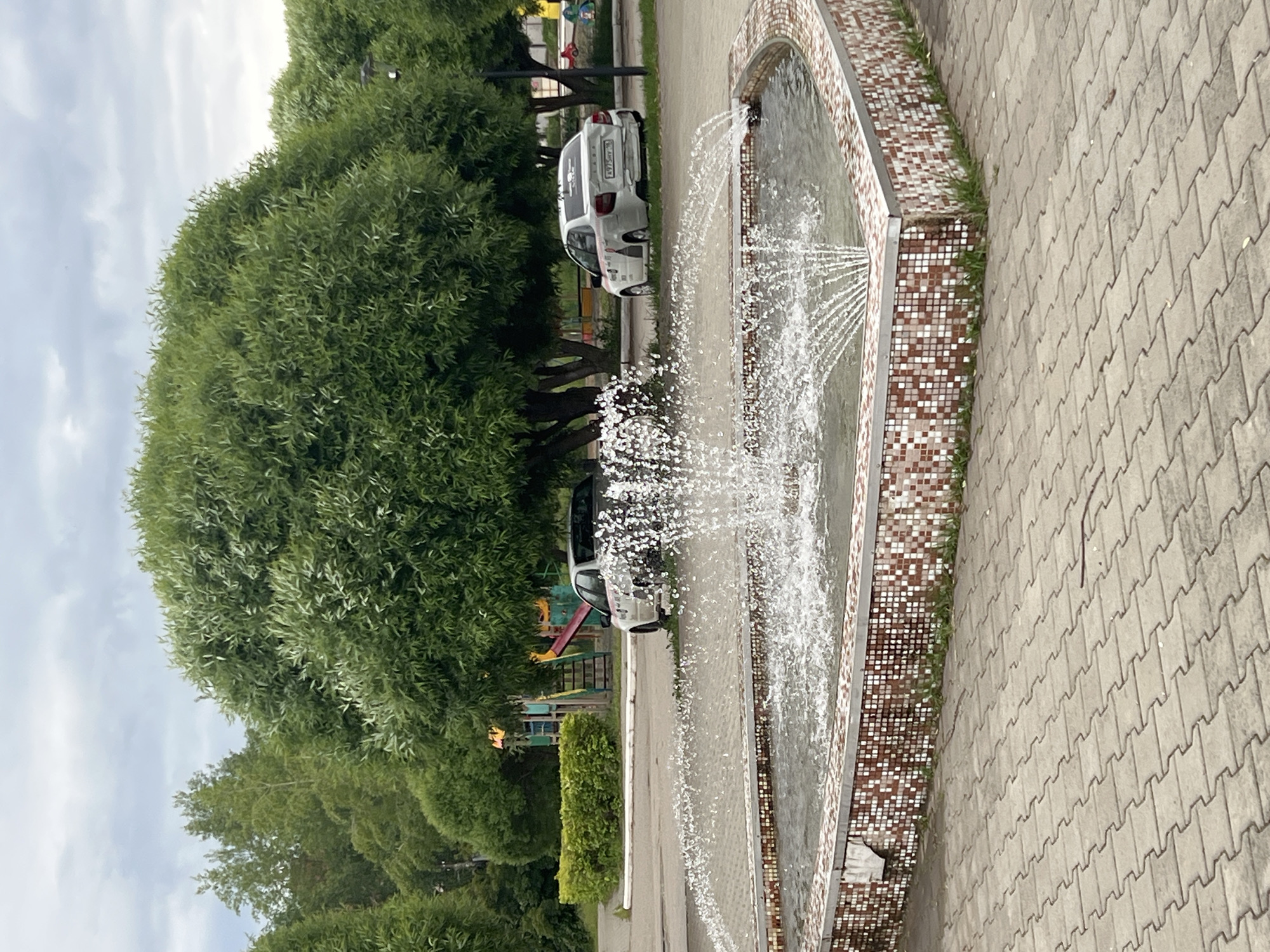 The image depicts an outdoor public space in Chaikovsky, Russia, on an overcast day. In the foreground, a heart-shaped fountain constructed from small, multi-toned mosaic tiles (white, brown, reddish-brown) actively sprays water from multiple jets into its basin. The fountain is surrounded by light grey interlocking concrete pavers, arranged in a herringbone pattern. Small patches of grass are visible at the base of the fountain's tiled wall.
In the midground, beyond the fountain, dense green deciduous trees dominate the scene. Two white sedan cars are parked on a grassy area beneath these trees. The car on the right is a white Lada Granta sedan, exhibiting significant damage to its rear bumper and trunk lid. Its Russian license plate, `A975MY 59`, is clearly legible. Faint decals are present on its rear passenger side. The car on the left, also a white sedan, is partially obscured by trees and the fountain's water spray.
The background reveals a children's playground structure with colorful elements, including red and yellow slides, situated behind the trees on the left side. A trimmed green hedge runs along the edge of a paved area near the playground. The sky above is uniformly overcast with grey clouds. No individuals are present within the frame.
