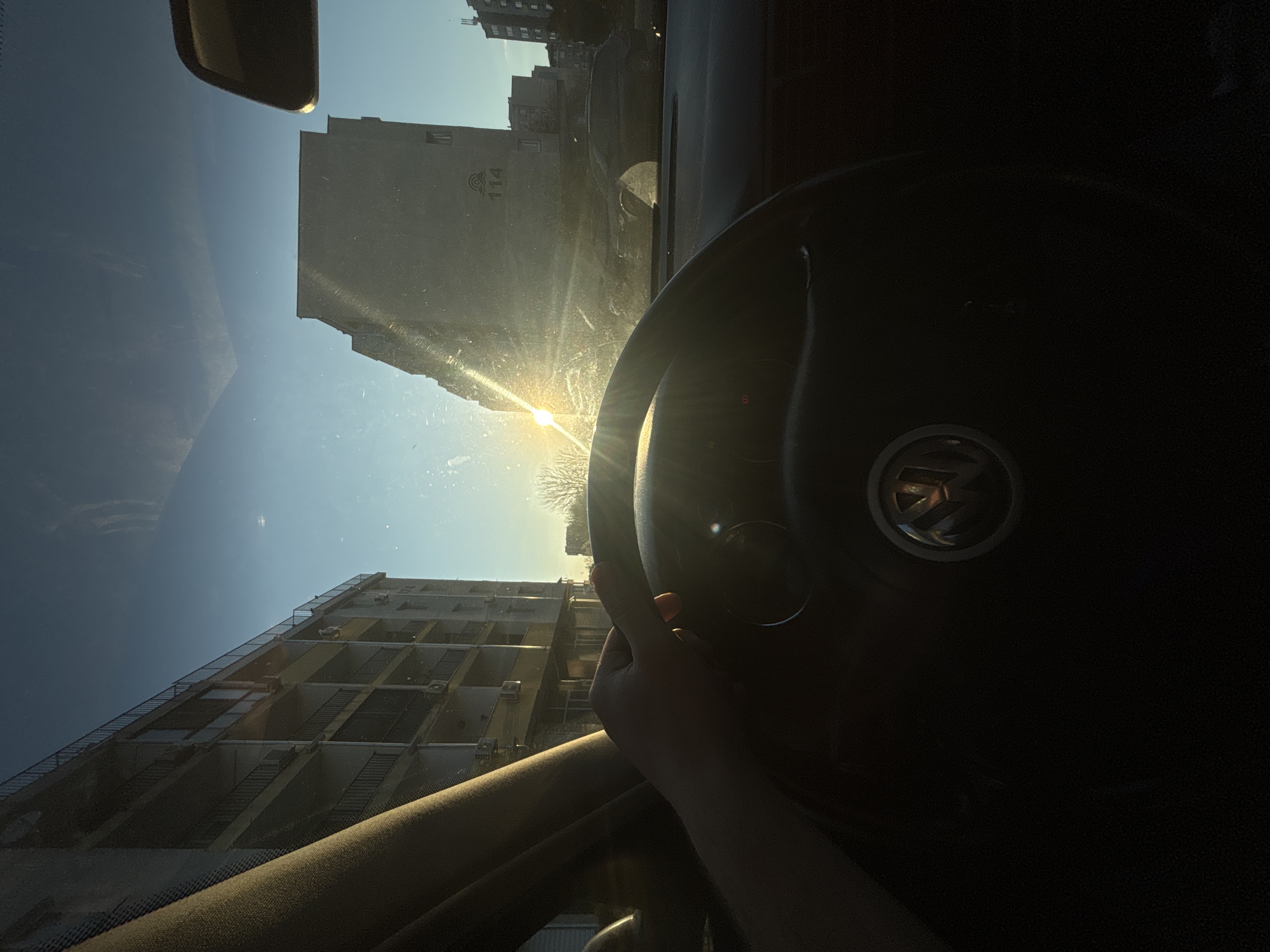 The photograph, taken from inside a car, shows a view of the exterior through the windshield. The sun is shining brightly, creating glare. In the foreground, a hand is visible on the steering wheel, which has a Volkswagen logo. The scene outside includes buildings, reflecting sunlight. One building bears the number "114." Based on this detail and architectural style, this image was likely taken in Burgas, Bulgaria.