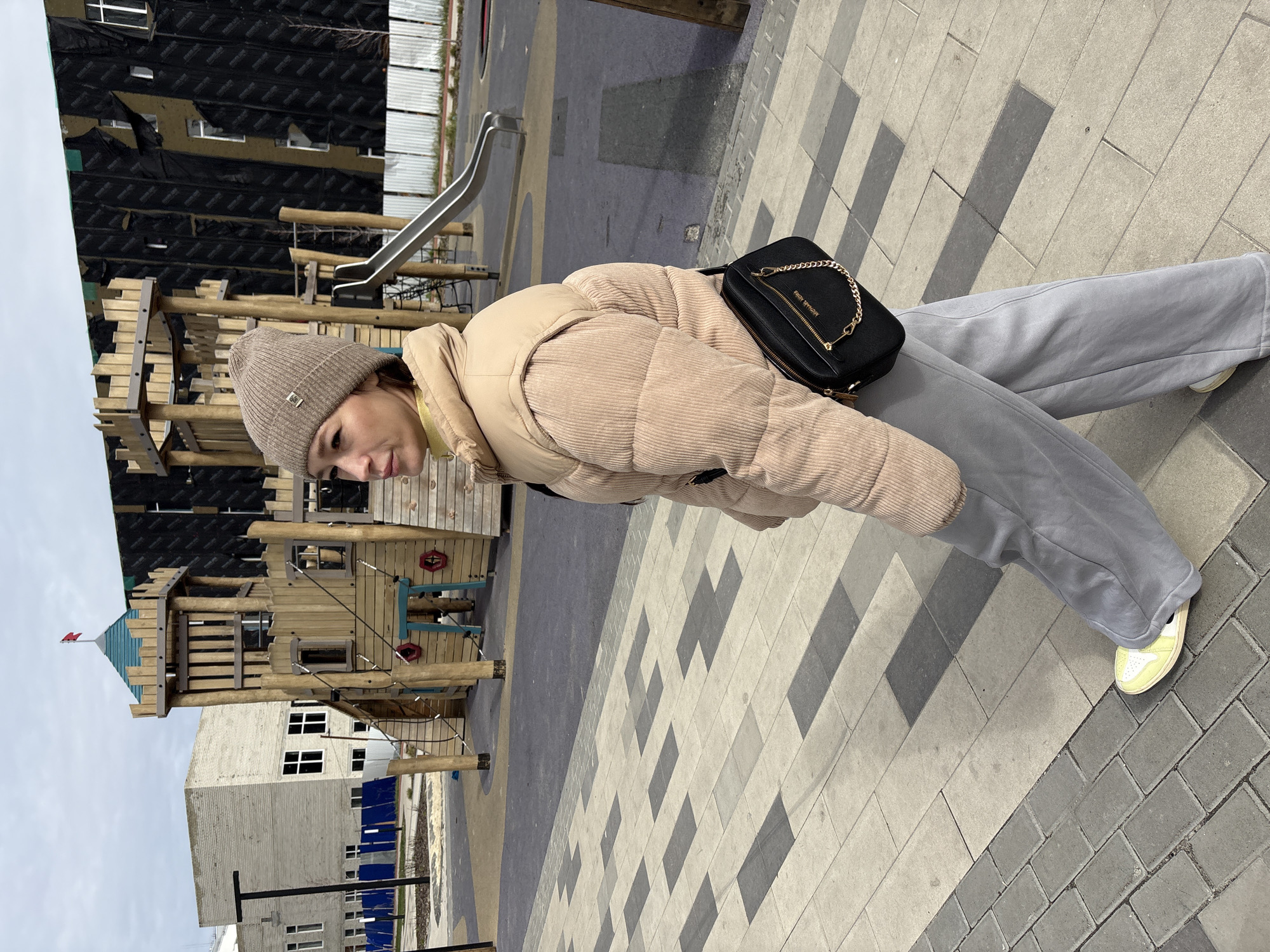 A woman stands on a paved surface in Ulyanovsk, Russia, next to a playground. She wears a beige knit hat, beige corduroy jacket, gray sweatpants, and yellow sneakers. She carries a black crossbody bag with a gold chain. Behind her is a wooden playground structure, a slide, and a building with visible black construction materials on its facade. The sky is overcast.