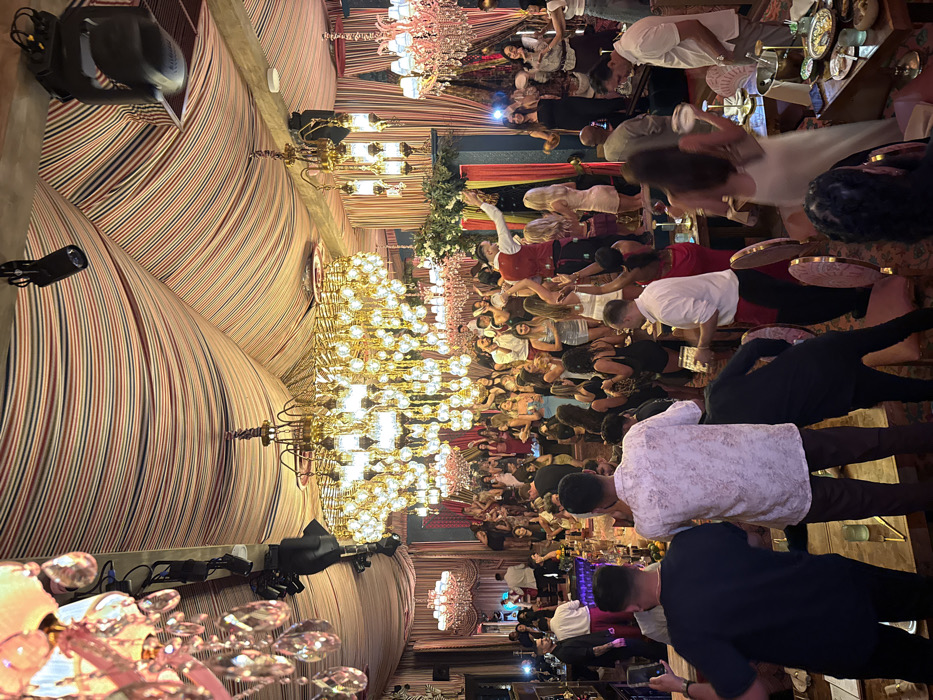 This image captures a lively indoor event at a restaurant or club, strongly resembling **Marion Miami** in the Brickell neighborhood of Miami, Florida, United States.
The scene depicts a large gathering of approximately 50-70 individuals within an elaborately decorated space. The ceiling is draped with red, white, and blue striped fabric, creating a tent-like canopy from which numerous ornate chandeliers hang. A prominent, multi-tiered golden chandelier with numerous glowing bulbs dominates the central overhead space, flanked by smaller, pink-hued crystal chandeliers and black stage spotlights.
The patrons, primarily adults in their 20s to 40s, are dressed in upscale casual to semi-formal attire, with women in various dresses and men in shirts and trousers. Many individuals are standing, dancing, or gesturing with raised hands, indicating a festive atmosphere. A male entertainer, wearing a red vest over a white shirt, stands on a small, fabric-decorated raised platform in the mid-ground, actively performing with outstretched arms. Other guests are observed socializing and holding drinks. In the foreground, three men stand with their backs to the camera, one on the far left holding a smartphone as if recording, positioned near a bar counter or high-top table. Tables with drinks and some food items are scattered throughout the venue.
