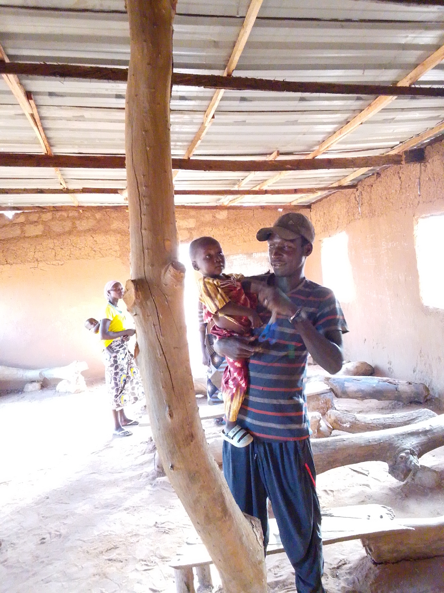 A man stands inside a building in Mulke Barikin Sayejo, Nigeria. He wears a cap, striped shirt, and blue pants and holds a child in his arms. Other people stand in the background. A large wooden support column stands prominently in the foreground, and logs or wooden beams are visible on the ground. The building appears to have walls constructed from earthen material and a corrugated metal roof supported by wooden beams. Windows are cut into the earthen walls. The scene appears to be sparsely furnished, with a sandy ground surface.
