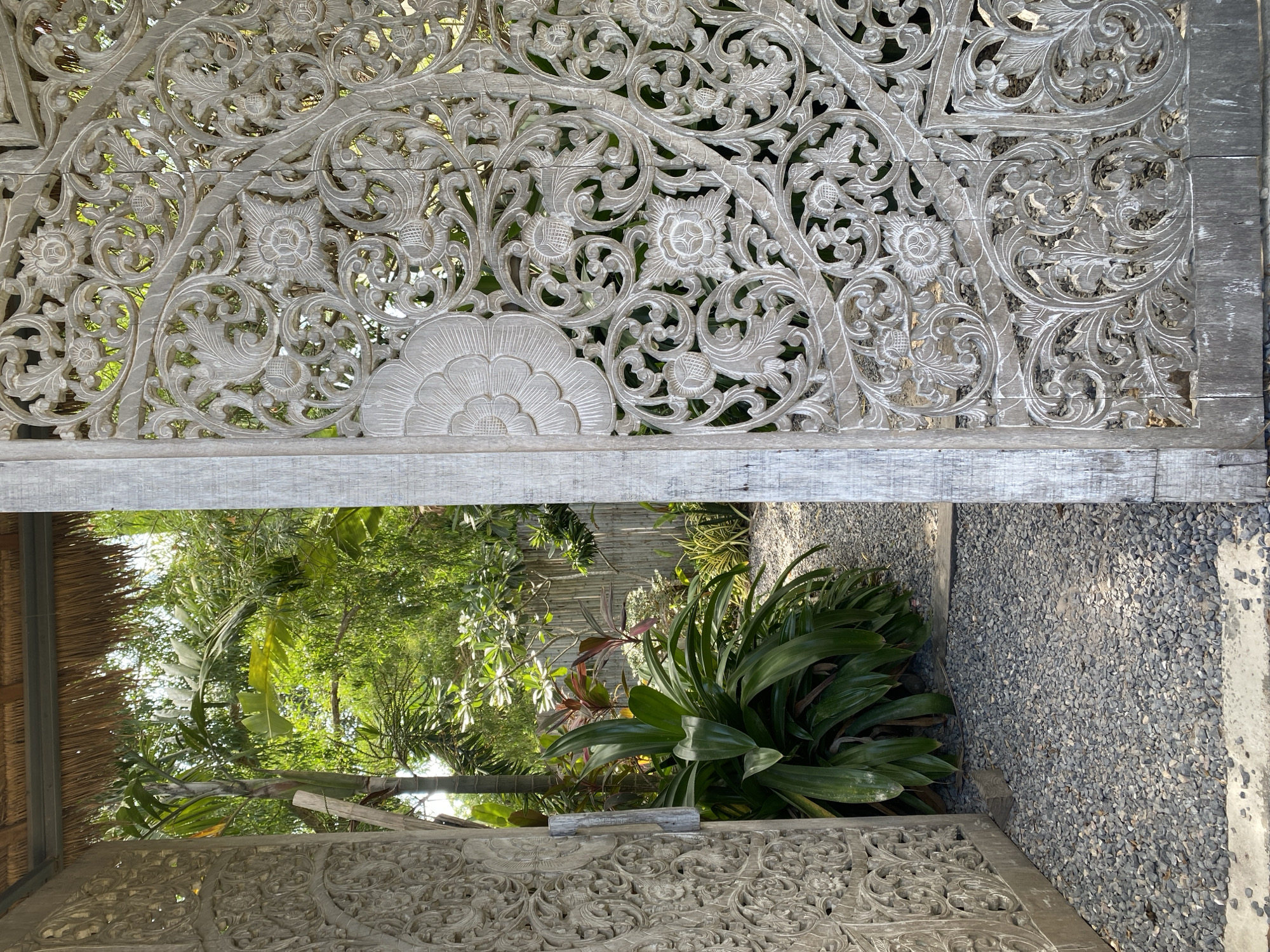 The image presents a detailed view looking through an elaborately carved wooden screen or gate into a lush tropical garden. The screen, made of light-colored, weathered wood, is adorned with intricate floral and organic patterns, including stylized flowers, swirling vines, and detailed leaves, indicating traditional craftsmanship, possibly of Balinese or Thai origin. The wood's slightly aged or whitewashed finish accentuates the relief of the carvings.
Beyond the screen's openwork, a vibrant natural scene unfolds, dominated by dense tropical foliage in various shades of green. Large-leafed plants, possibly banana leaves, mix with smaller bushes and other verdant growth, suggesting a thriving garden environment. To the right, a ground area covered with small, grey gravel is visible. A partial view of a traditional thatch roof can be seen on the far left, hinting at the architectural style of nearby structures.
The scene is illuminated by bright, natural daylight, implying clear weather, likely morning or late afternoon, with sunlight highlighting the leaves. There are no people or specific activities taking place, contributing to a tranquil atmosphere. The setting, with its tropical vegetation and intricate wood carving, aligns well with the provided location context of Baan Sri Thanu, Thailand. No visible text is present in the media file.