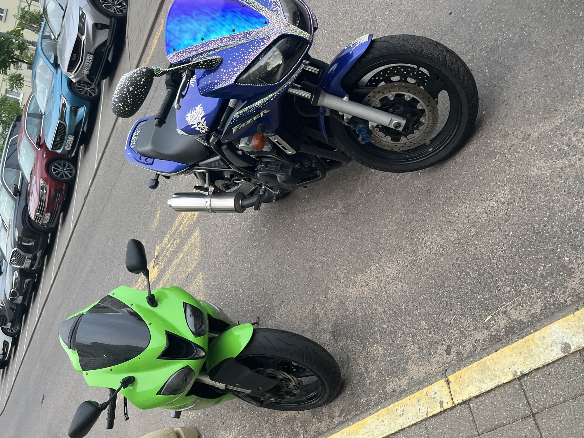 Two motorcycles are parked on an asphalt street next to a sidewalk with a yellow curb. The motorcycle on the left is bright green with a black windshield and black mirrors. The motorcycle on the right is blue and decorated with rhinestones on its mirrors, front fairing, and body. It also has a chrome exhaust pipe. In the background, several cars are parked, including a red Volkswagen, a blue Geely, and a silver Geely. The scene appears to be a street in Minsk, Belarus, based on the vehicle license plates which are characteristic of Belarus.