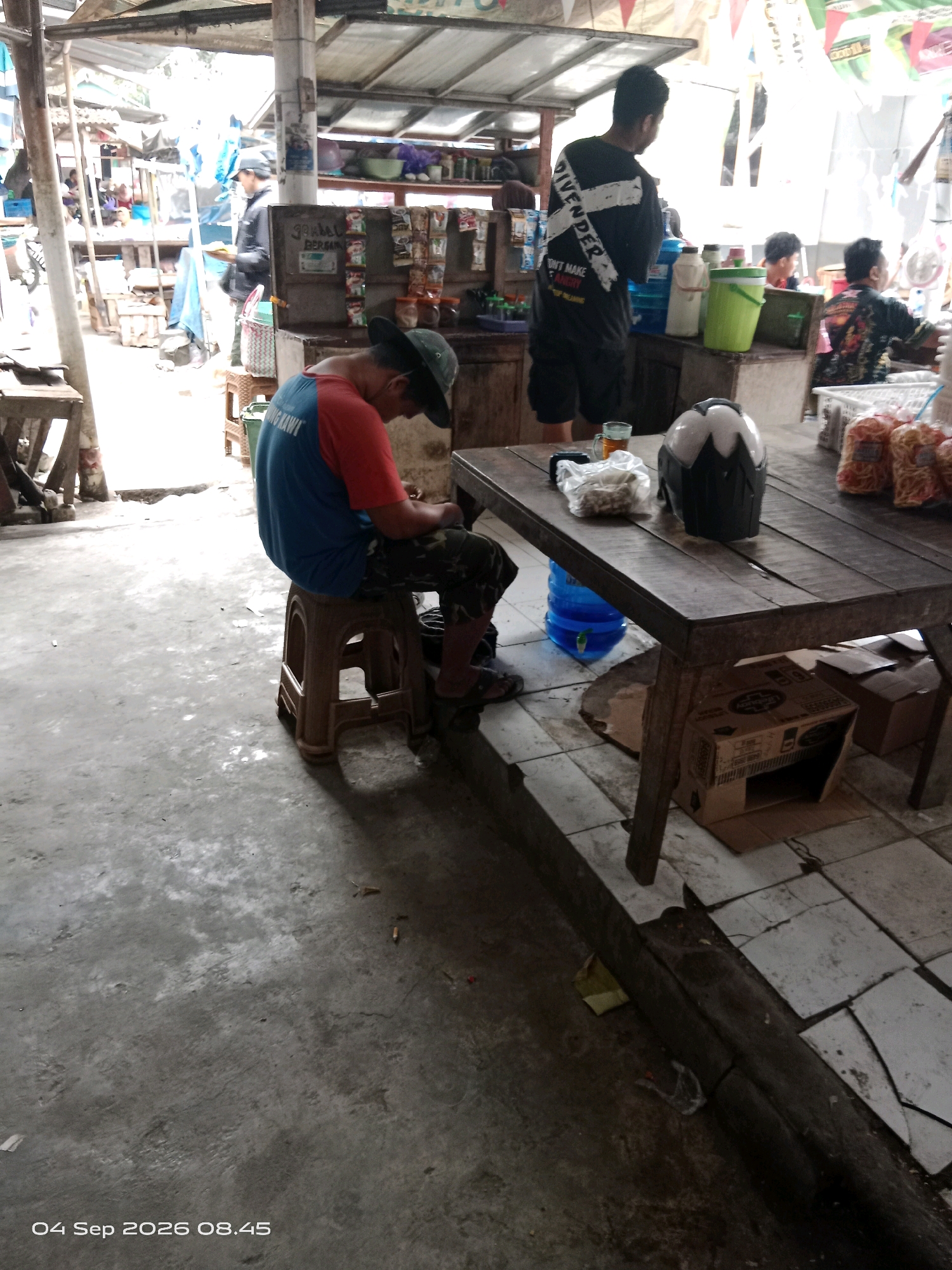 In Kalipurwo, Indonesia, a man wearing a blue and red t-shirt and a dark green hat sits on a plastic stool at a market stall. He is looking down at his hands. In front of him is a wooden table with a helmet, a plastic bag filled with snacks, a glass of tea, and a blue water dispenser. Behind the man, another person wearing a black t-shirt with white writing stands near a shelf displaying various products. The market appears to be outdoors, with a corrugated metal roof overhead. The ground is concrete with some scattered debris.