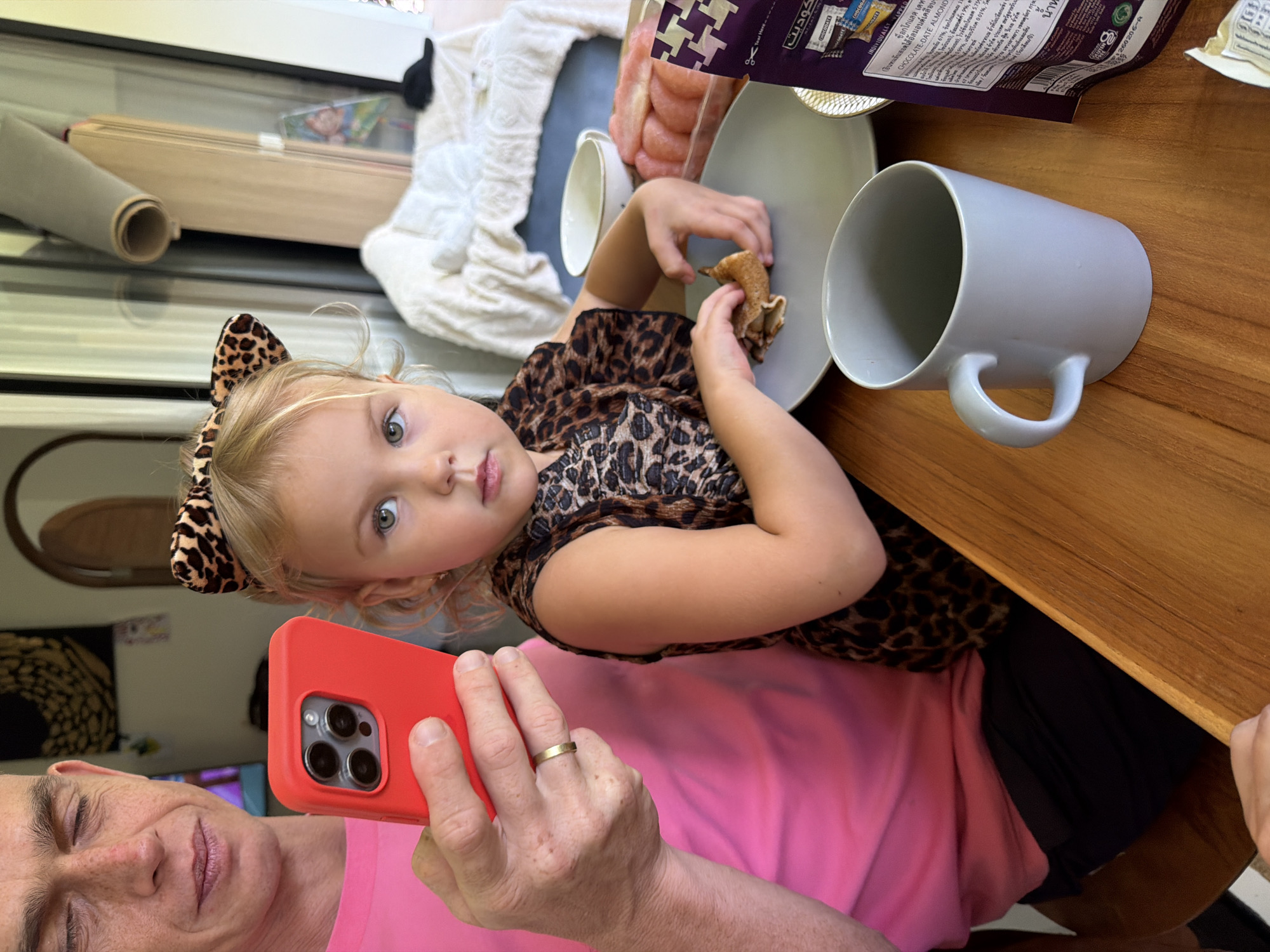 A person, presumably male based on the hands and facial features, is seated at a wooden table, holding a red smartphone in his left hand. He is wearing a bright pink t-shirt and dark pants. His eyes are closed, and he appears to be looking at the phone. Seated next to him, also at the table, is a young girl. She has blonde hair styled with a leopard-print headband. She is wearing a leopard-print top. The girl is holding a piece of food, possibly a pancake or crepe, in her hands and is about to eat it. In front of her is a gray plate with the food and a gray mug. To her right, there is a clear plastic container filled with sliced pink fruit, likely grapefruit or pomelo, and a purple bag with some text on it. The background shows a room with a rolled-up mat on a shelf and furniture. The scene is likely indoors, possibly in a home or restaurant in Koh Phangan, Thailand, given the presence of the rolled mat which is often associated with yoga or meditation retreats popular in the area, and the tropical lighting.