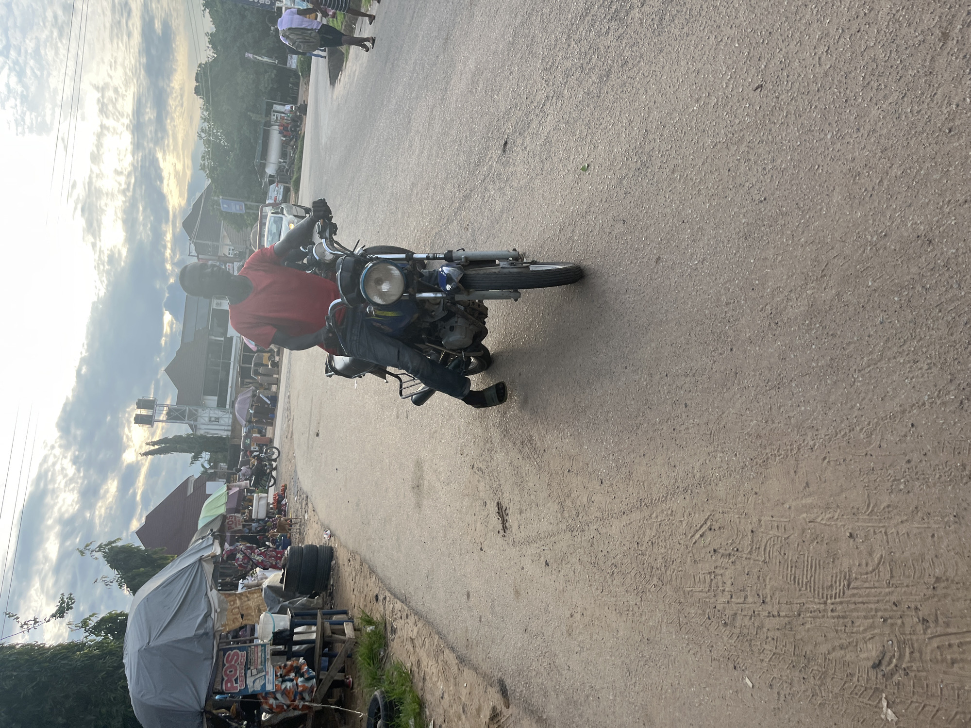 The image depicts a roadside scene, consistent with a commercial or semi-urban area in Nigeria. The specific location within the city of Abuja cannot be determined from the visual information provided.
In the foreground, a man, appearing to be of African descent, wears a red short-sleeved shirt, dark blue trousers, and dark slides/sandals. He is seated on a dark-colored motorcycle, holding the handlebars, with his left foot on the ground. The motorcycle has a round headlight and visible engine components. The road surface in the immediate foreground is paved and grey, exhibiting a slightly uneven texture with scattered debris. Tire and footwear impressions are visible on the unpaved dirt shoulder to the left.
To the left of the motorcycle, a makeshift market area is visible. This area includes several stalls covered by large grey umbrellas. A prominent sign displaying "POS SERVICE" in red and white lettering is attached to one of these stalls. A wooden bench, a blue plastic drum, and stacked black tires are also present. Partially visible individuals are situated near these stalls.
The road extends into the midground, curving slightly to the right. On the far side of the road, several individuals are walking, including one person wearing a backpack and another in a striped garment. Further down the road, other vehicles, including cars and motorcycles, are discernable.
The background features various buildings with different roof types, including one prominent structure with a red tiled roof. Dense green foliage and trees are abundant. A tall, light-colored metal structure, possibly a water or communication tower, stands on the right side of the background. The sky is largely cloudy with patches of lighter areas, suggesting the sun is low on the horizon, creating soft, diffused illumination across the scene.